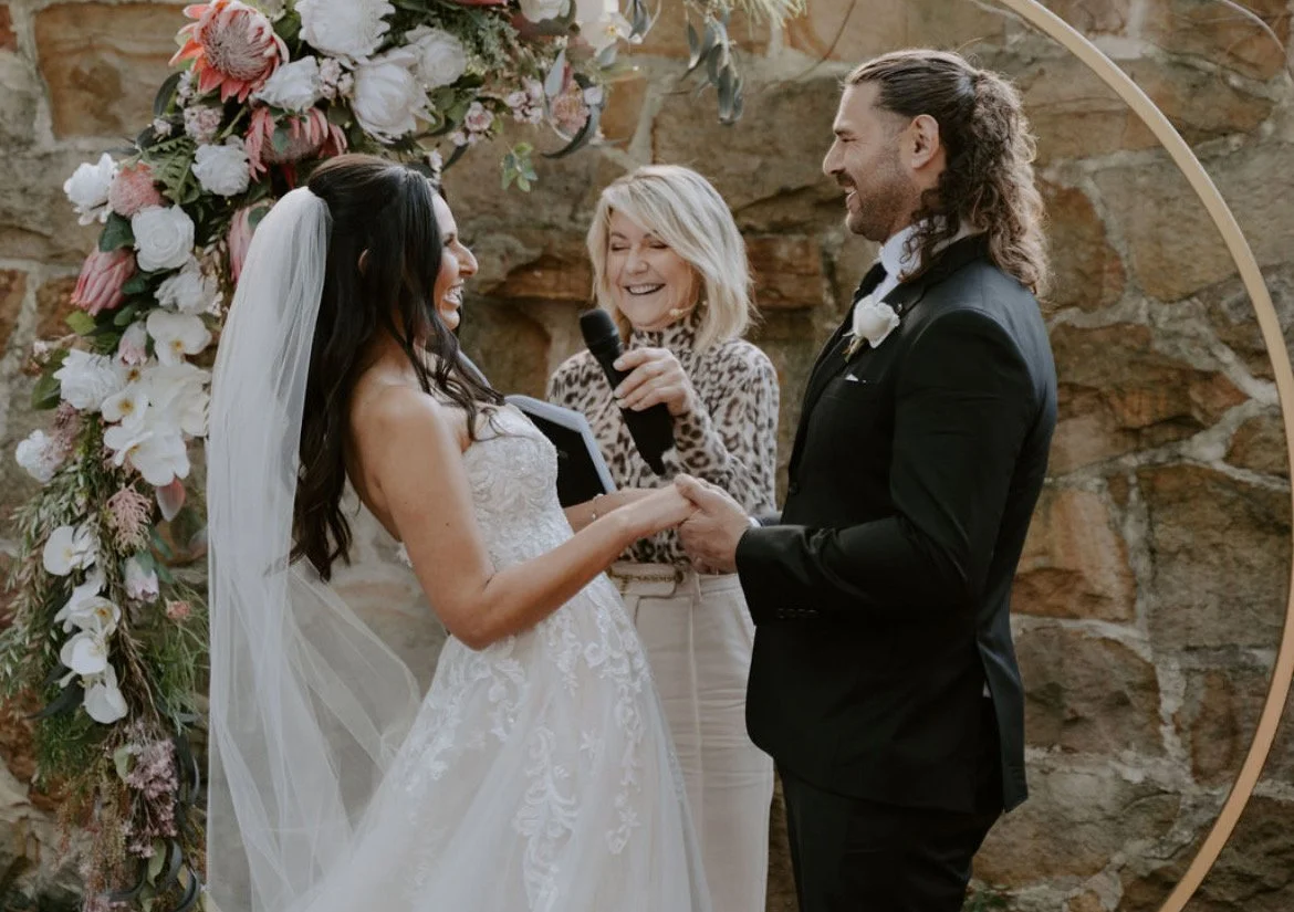 A wedding ceremony with a bride and groom holding hands and exchanging vows, standing in front of a floral arch, with their wedding celebrant Lisa speaking and smiling.