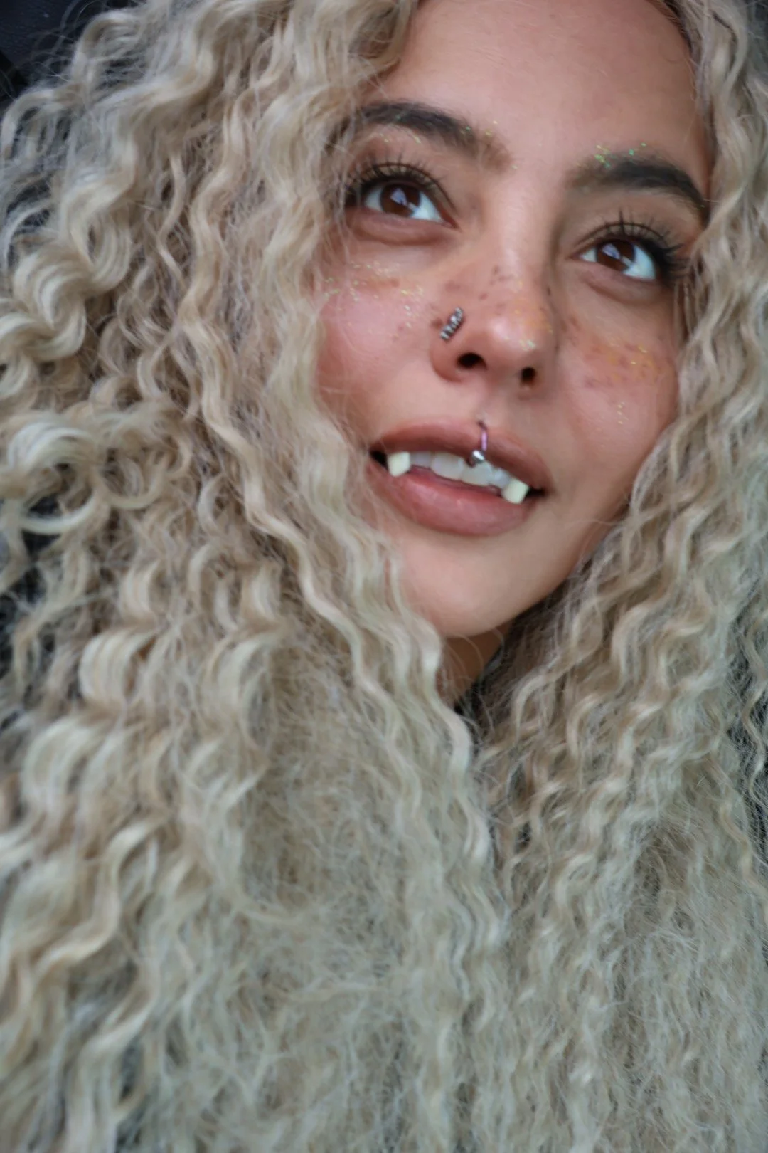 Close-up of a woman with curly blonde hair and multiple facial piercings, including a nose ring and a lip ring, wearing glitter on her face and looking upward.