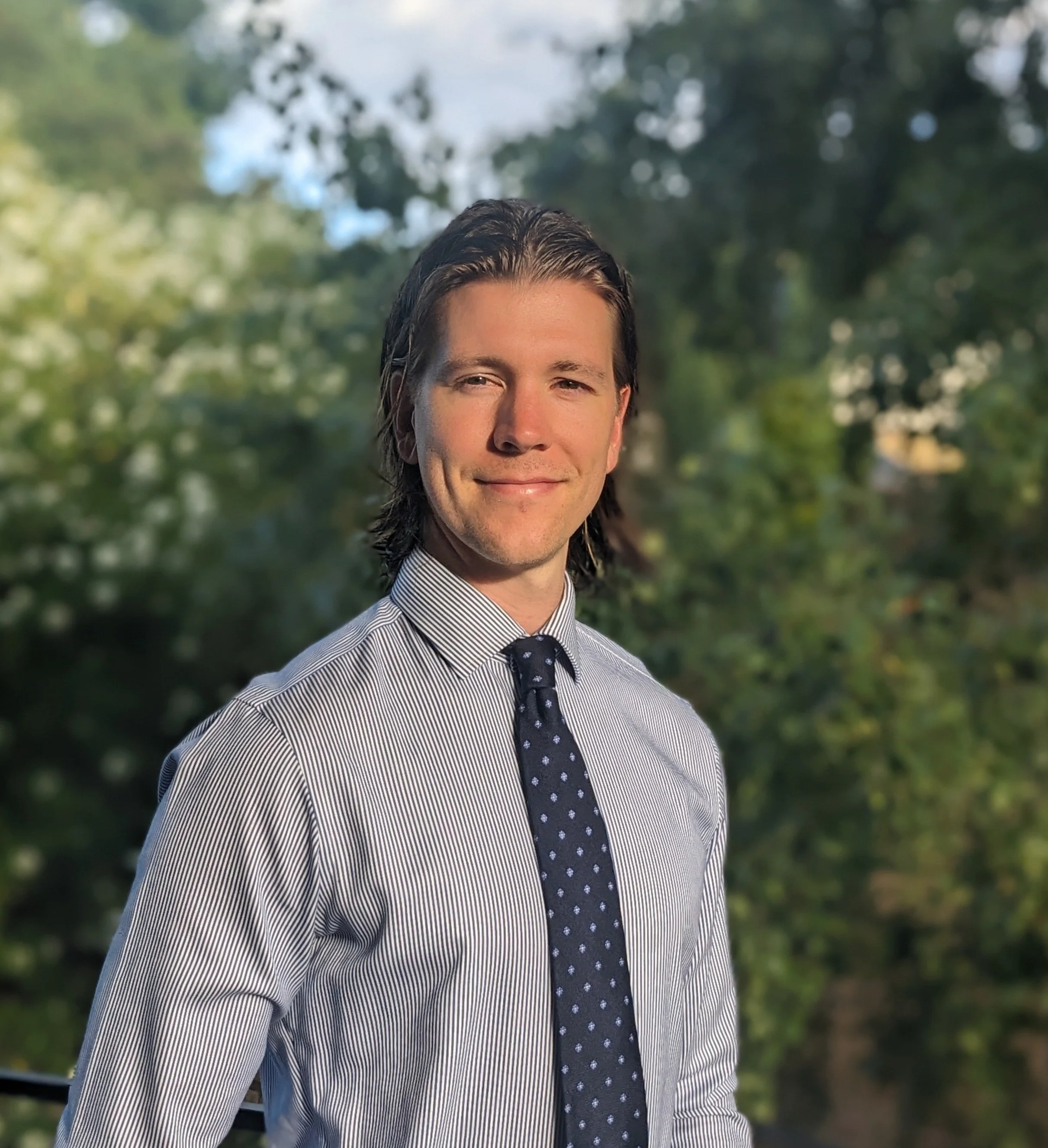 A man with shoulder-length dark brown hair, wearing a striped dress shirt and a navy tie with white polka dots, standing outdoors with a background of green trees and a partly cloudy sky.