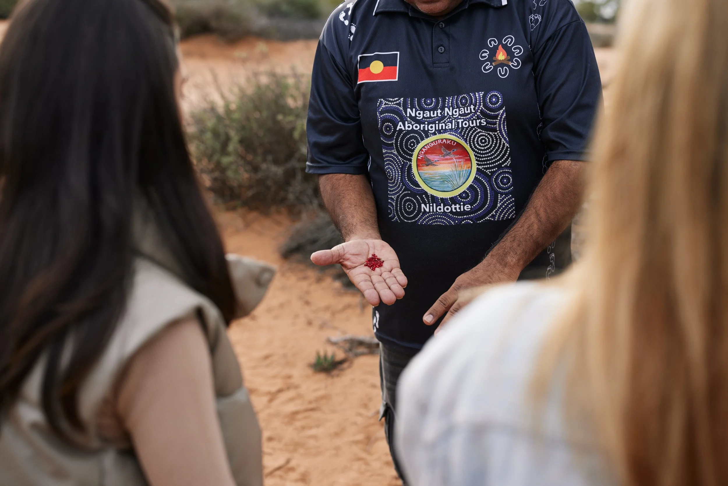 A person in a navy blue shirt with indigenous artwork and text holding a handful of red berries, showing them to two women with brown and blond hair, outdoors with desert vegetation and sand background.