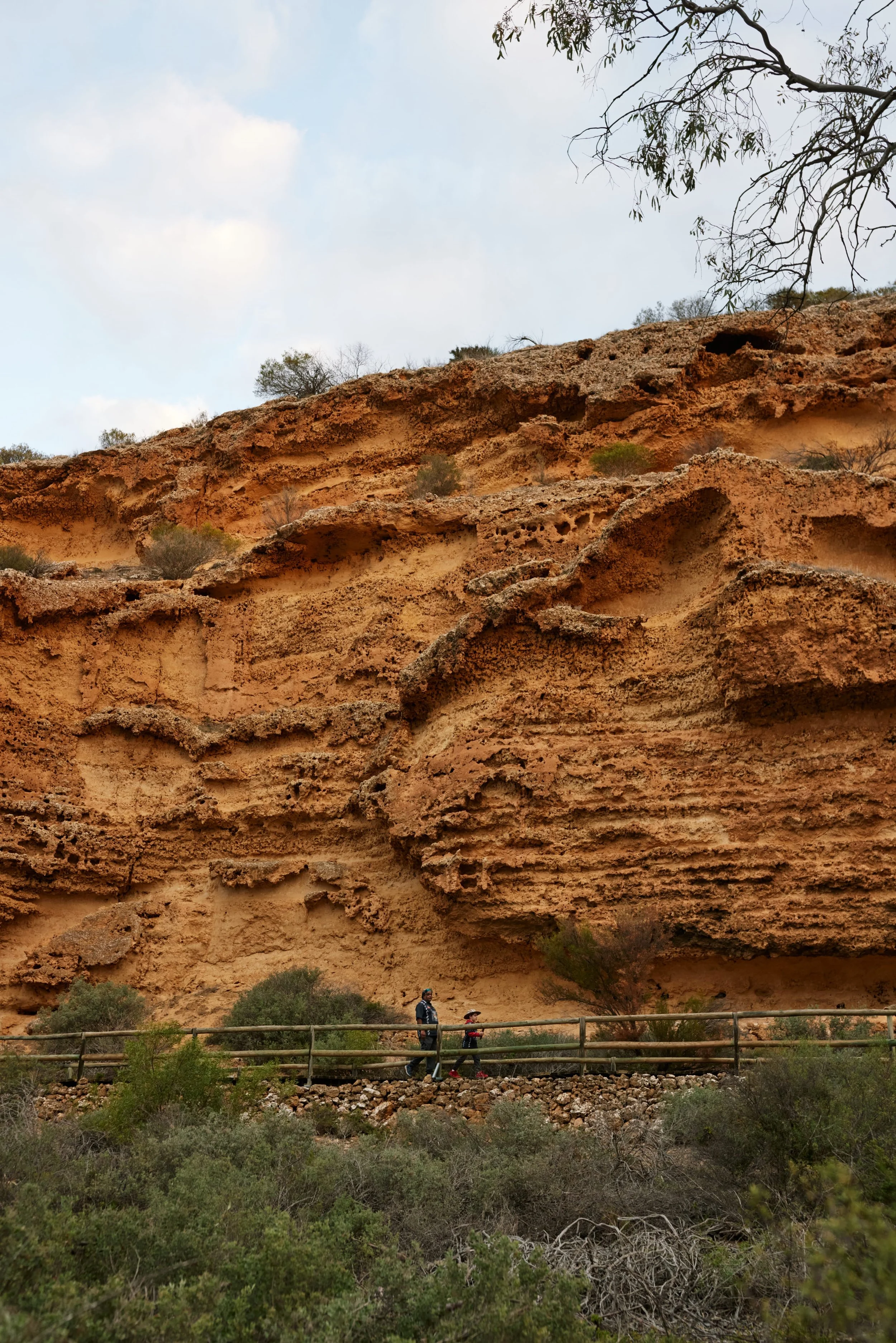 Two people walking on a trail with a wooden railing in front of a large, reddish-brown rock formation with sparse vegetation.
