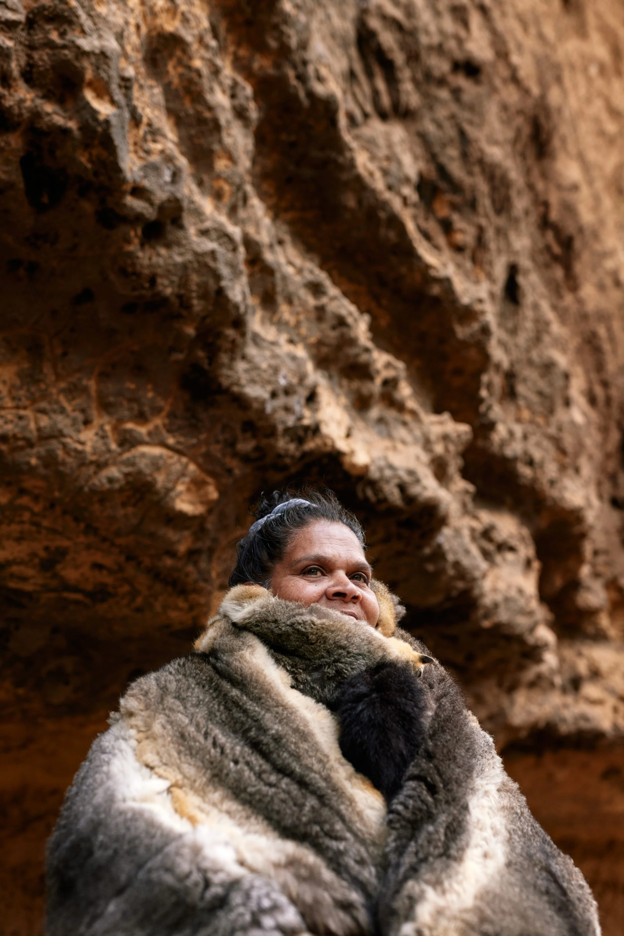 A woman wrapped in a thick fur coat, standing in front of a rocky cliff face.