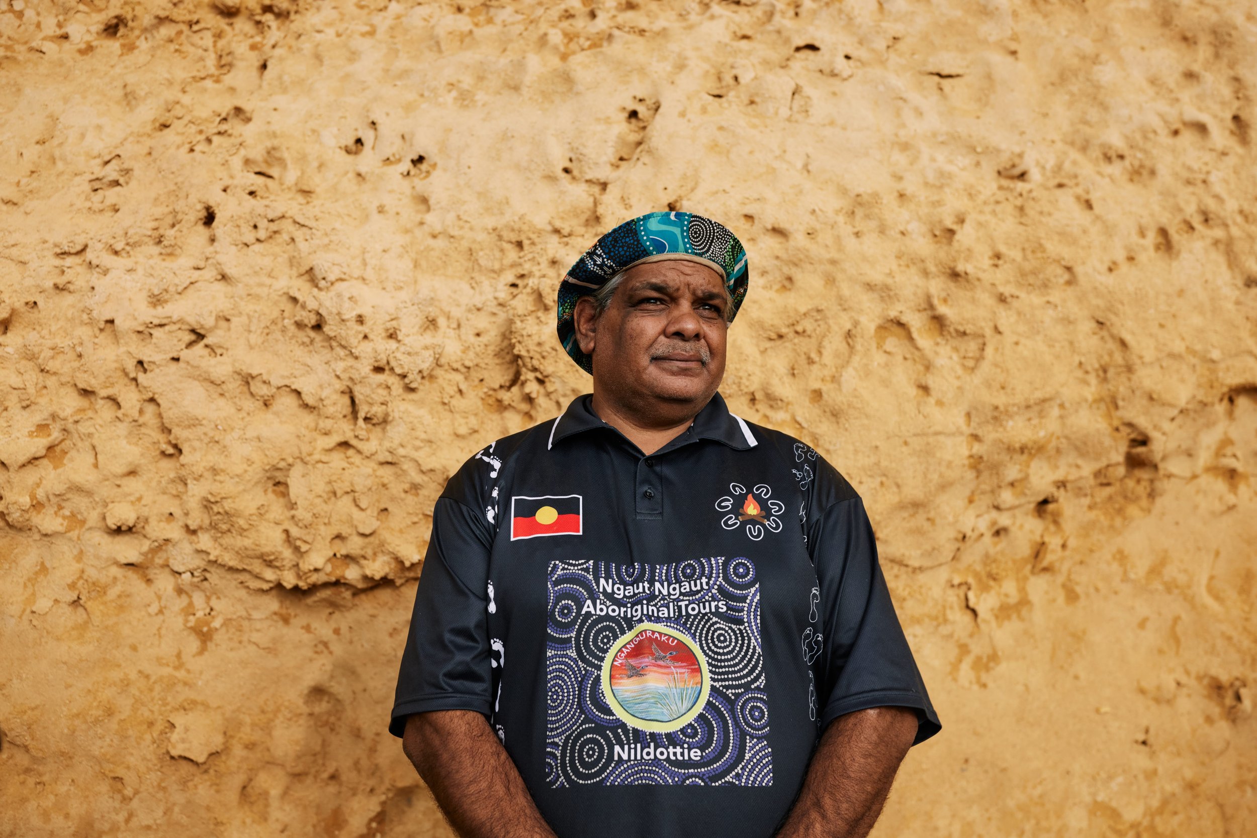 A man wearing a black shirt with indigenous artwork and a flag patch on the chest, and a patterned head covering, stands in front of a textured yellow wall, looking thoughtfully into the distance.
