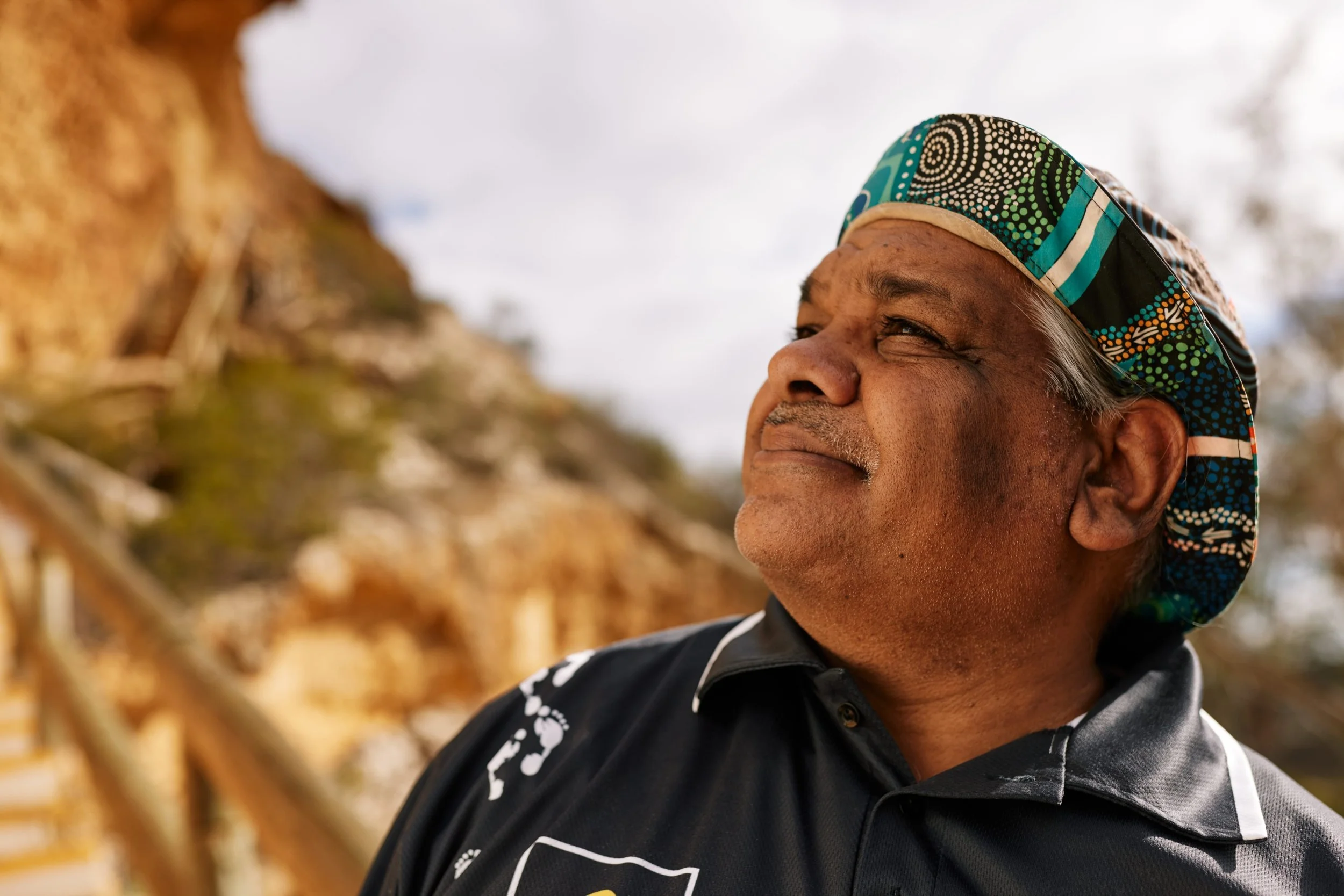 A middle-aged man in a colorful patterned hat and black sports shirt looking upwards outdoors with a blurred background of trees and a wooden staircase.