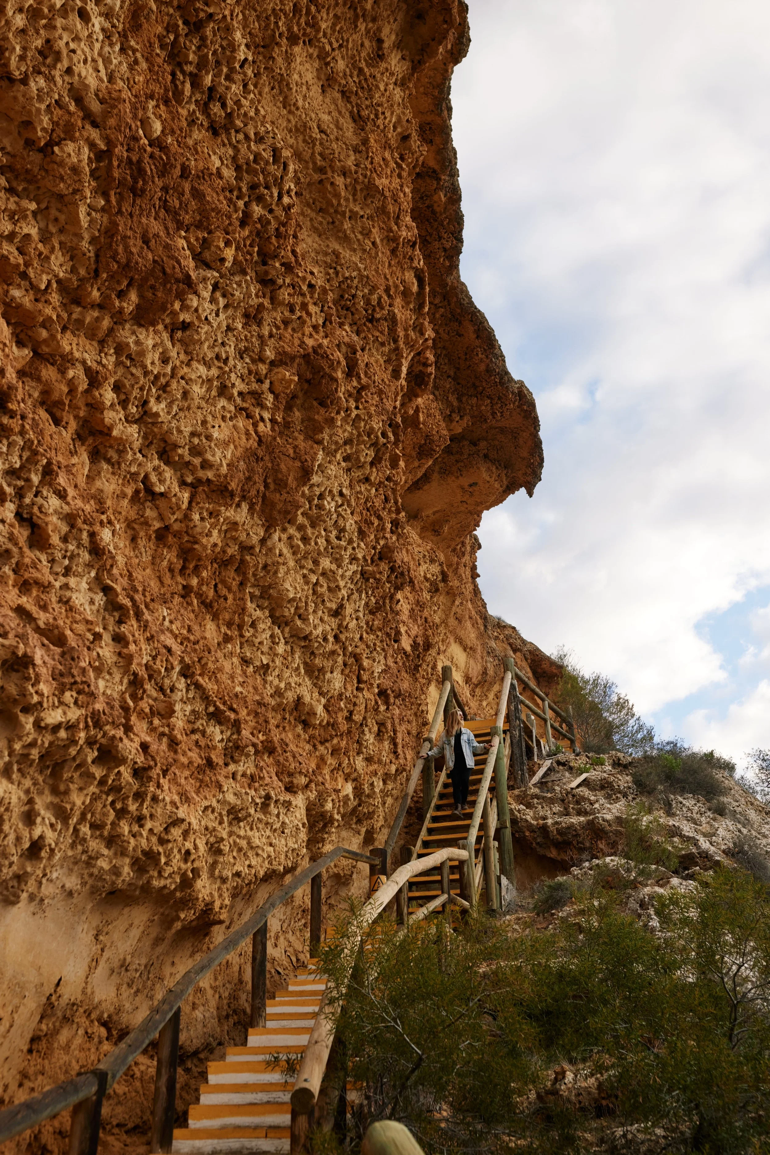 A person walking up a wooden staircase built along a tall, textured, reddish-brown rock formation outdoors.
