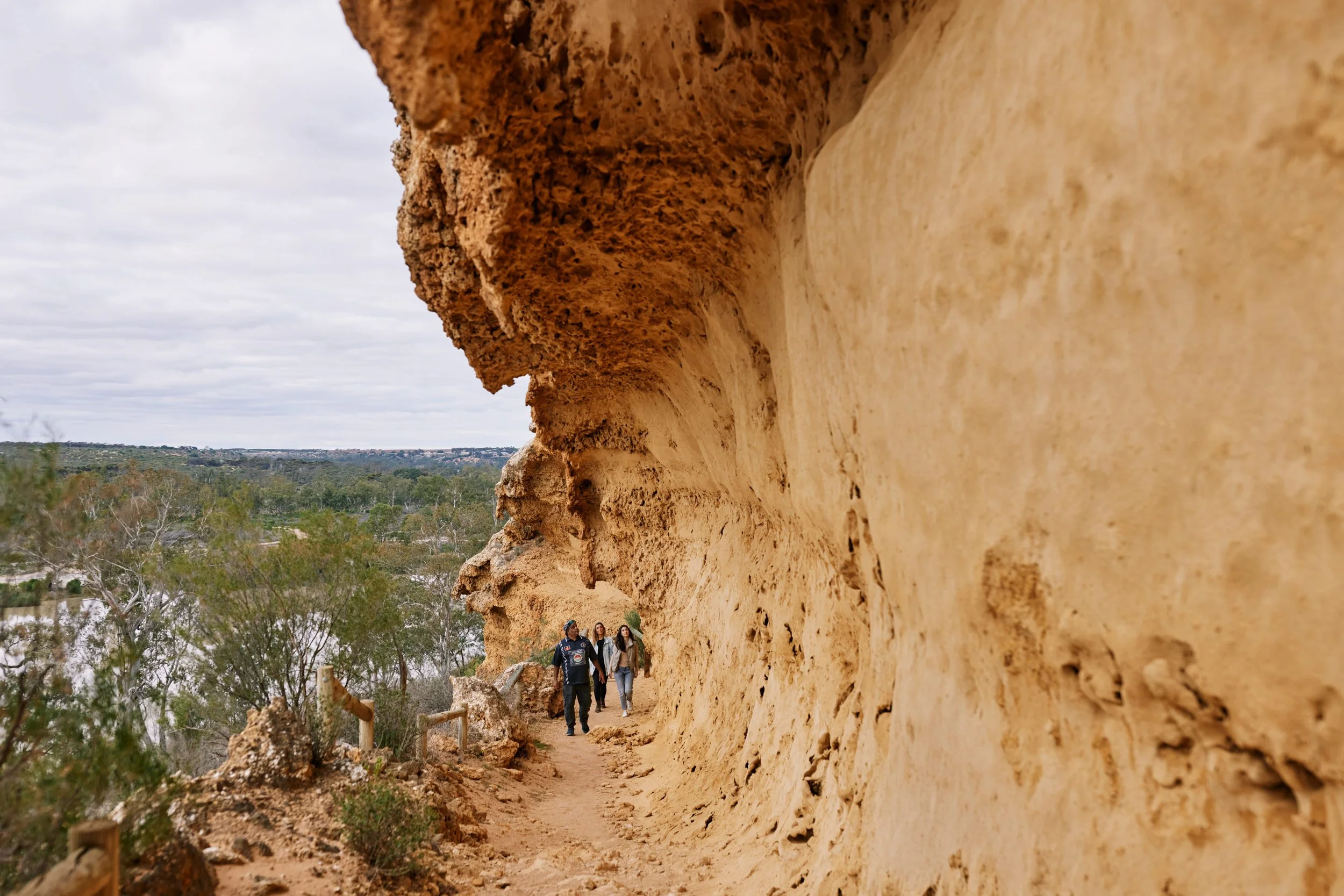 Three people walking along a narrow trail next to a large, overhanging sandstone rock formation in a natural outdoor setting with trees and cloudy sky in the background.