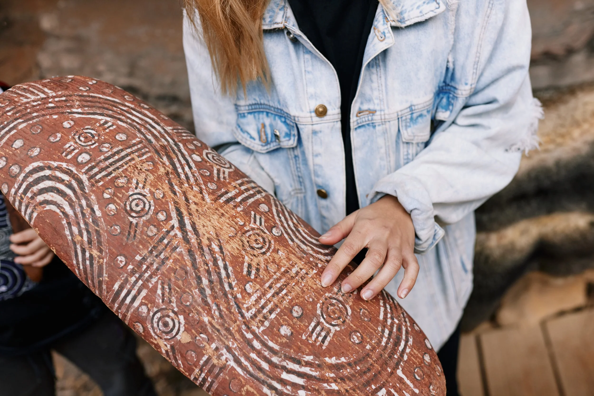 Person holding a circular object with intricate white tribal-like patterns on a red background, wearing a light denim jacket.