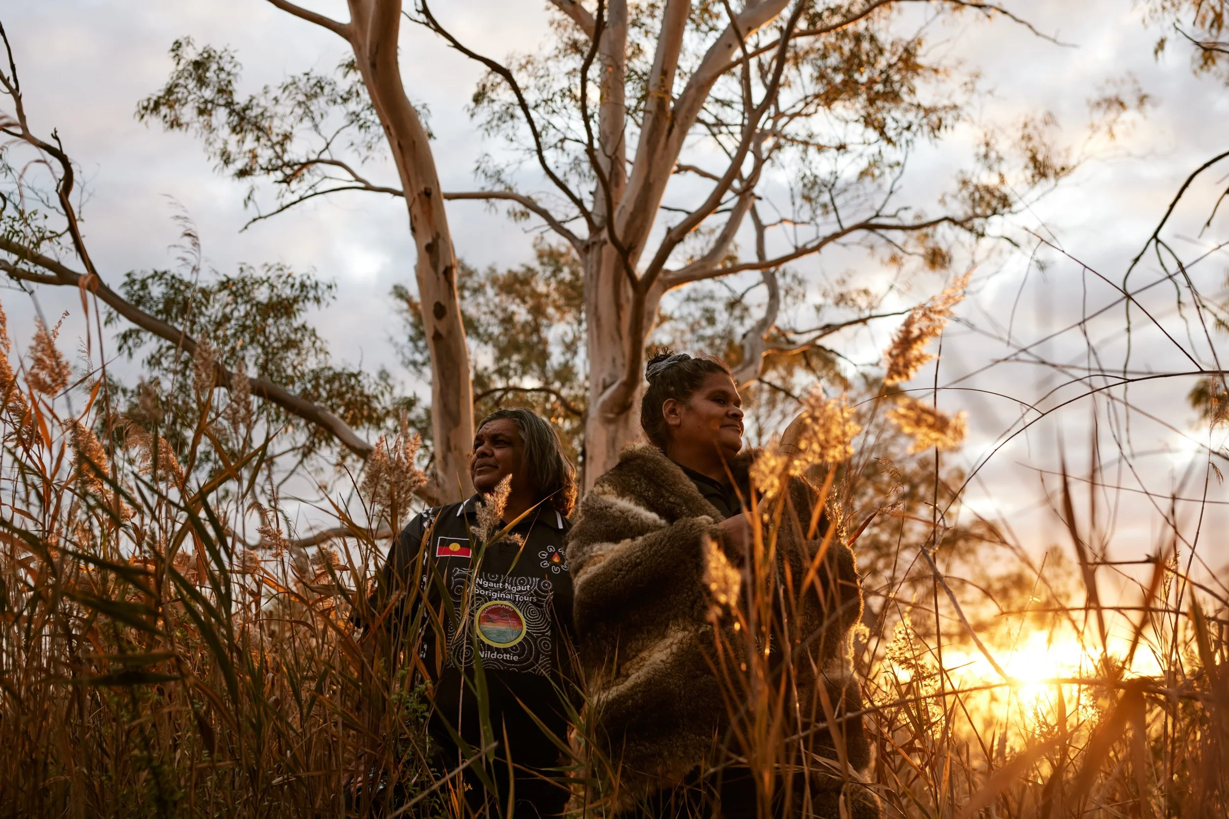Two Indigenous women standing in tall grass during sunset, with a large tree behind them, one wearing a fur coat and the other wearing a black jacket with patches.