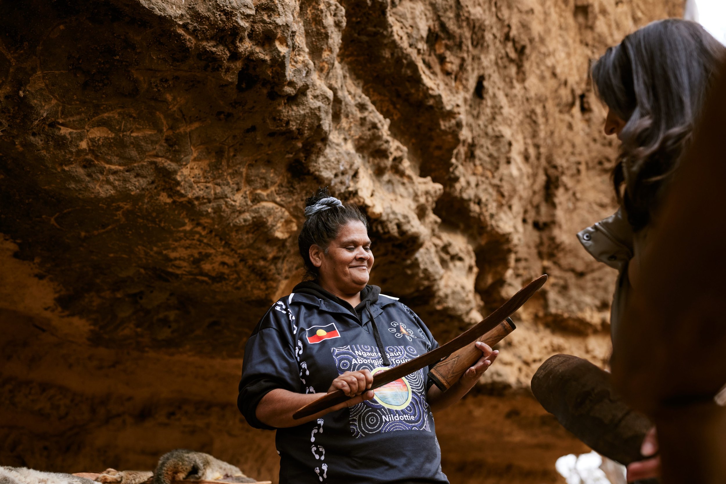 Indigenous woman holding traditional tool inside a rocky cave with another person partially visible.