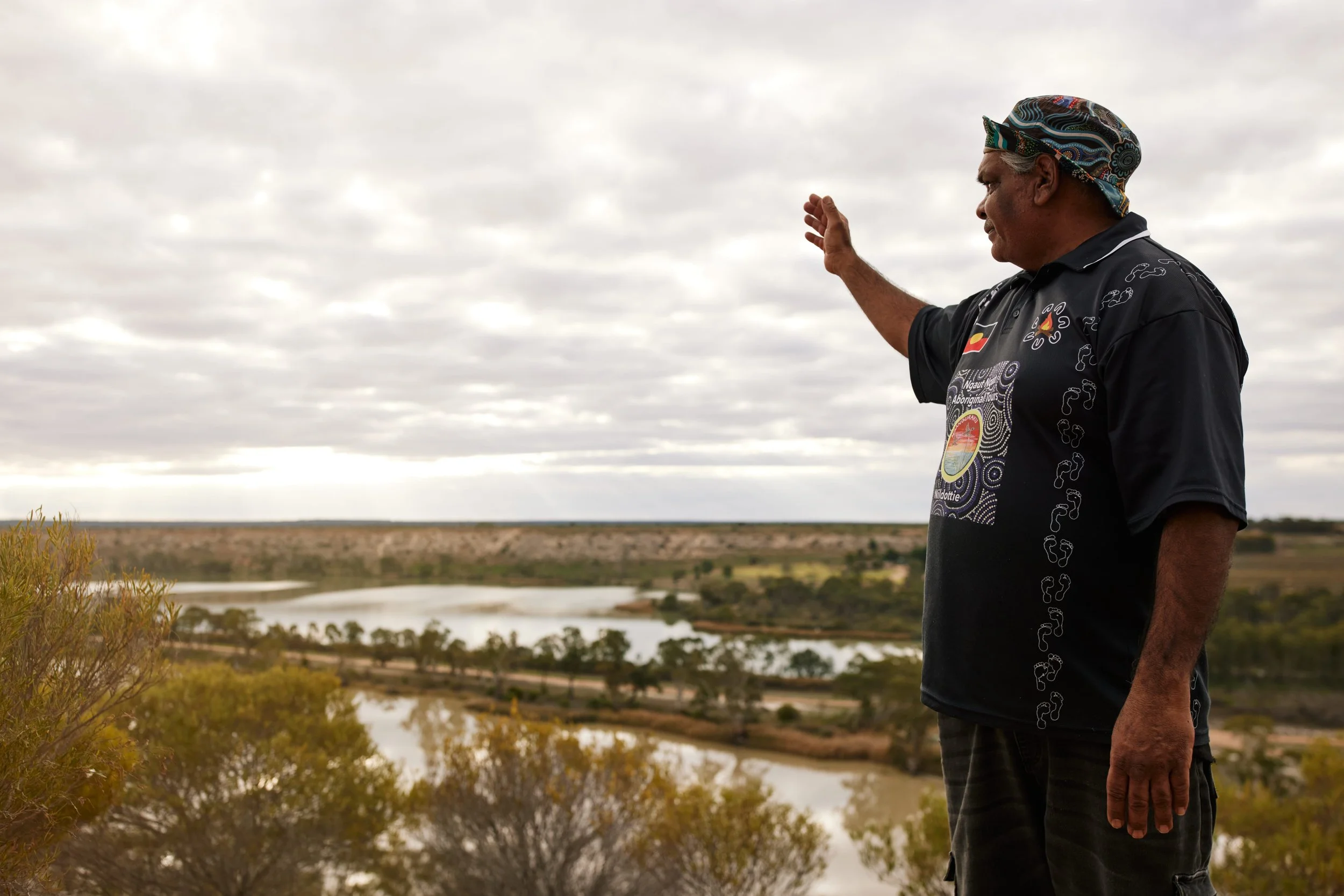 A man wearing a black shirt and patterned headband gestures with his right hand while standing outdoors near a body of water with trees and vegetation, under a cloudy sky.