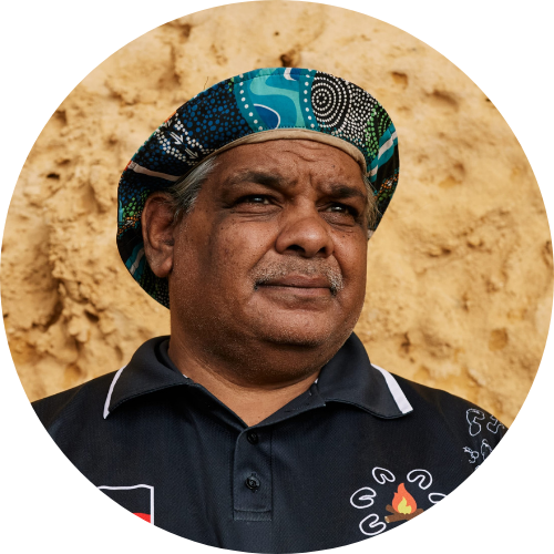 A man with dark skin wearing a black polo shirt and a colorful patterned hat, standing in front of a beige dirt wall.