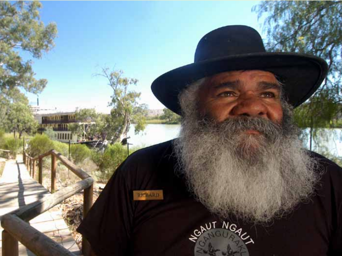 A man with a long gray beard wearing a black hat and black shirt standing outdoors near a river, with trees and a building in the background.