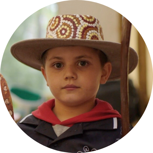 A young boy wearing a wide-brimmed hat with colorful circular patterns, a red hoodie, and a dark jacket, standing indoors.