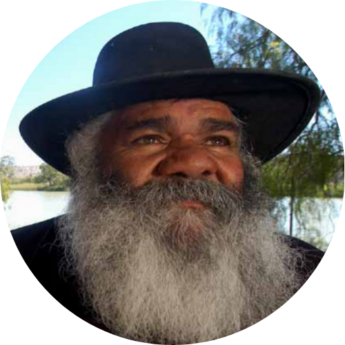 A man with a gray beard wearing a wide-brimmed black hat outdoors near a body of water with trees in the background.