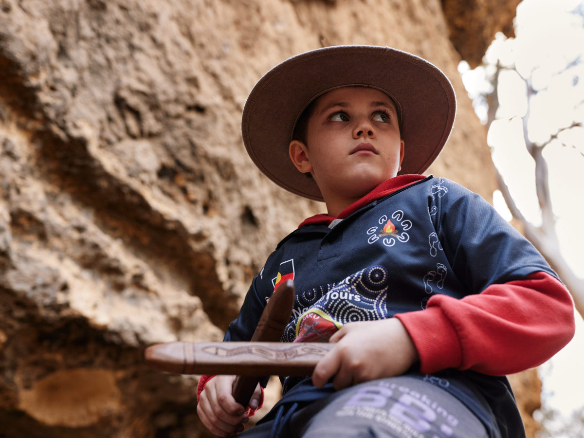 A boy wearing a wide-brimmed hat and a blue jacket with red sleeves, holding a wooden paddle, sitting outdoors near a rocky surface with trees in the background.