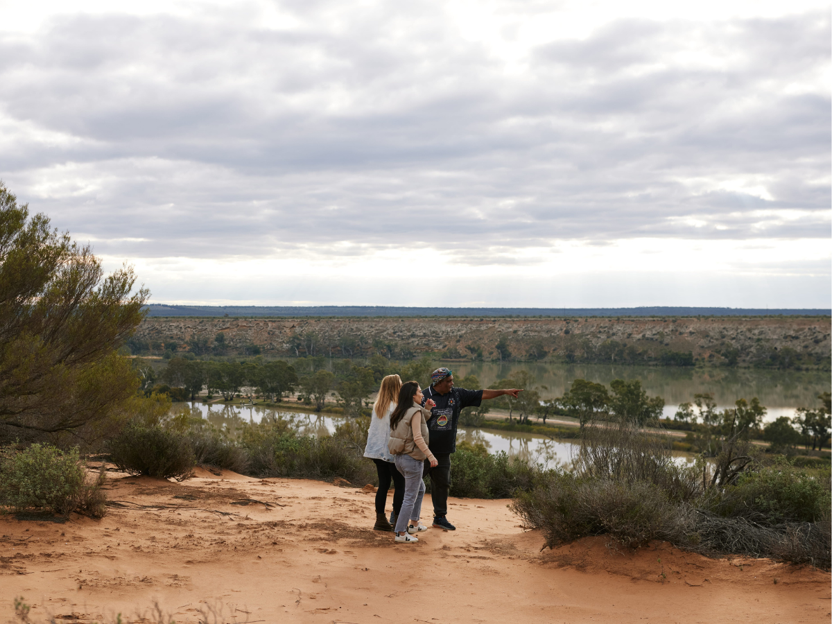 Group of three people standing on dirt trail overlooking a river landscape with trees and hills under cloudy sky.