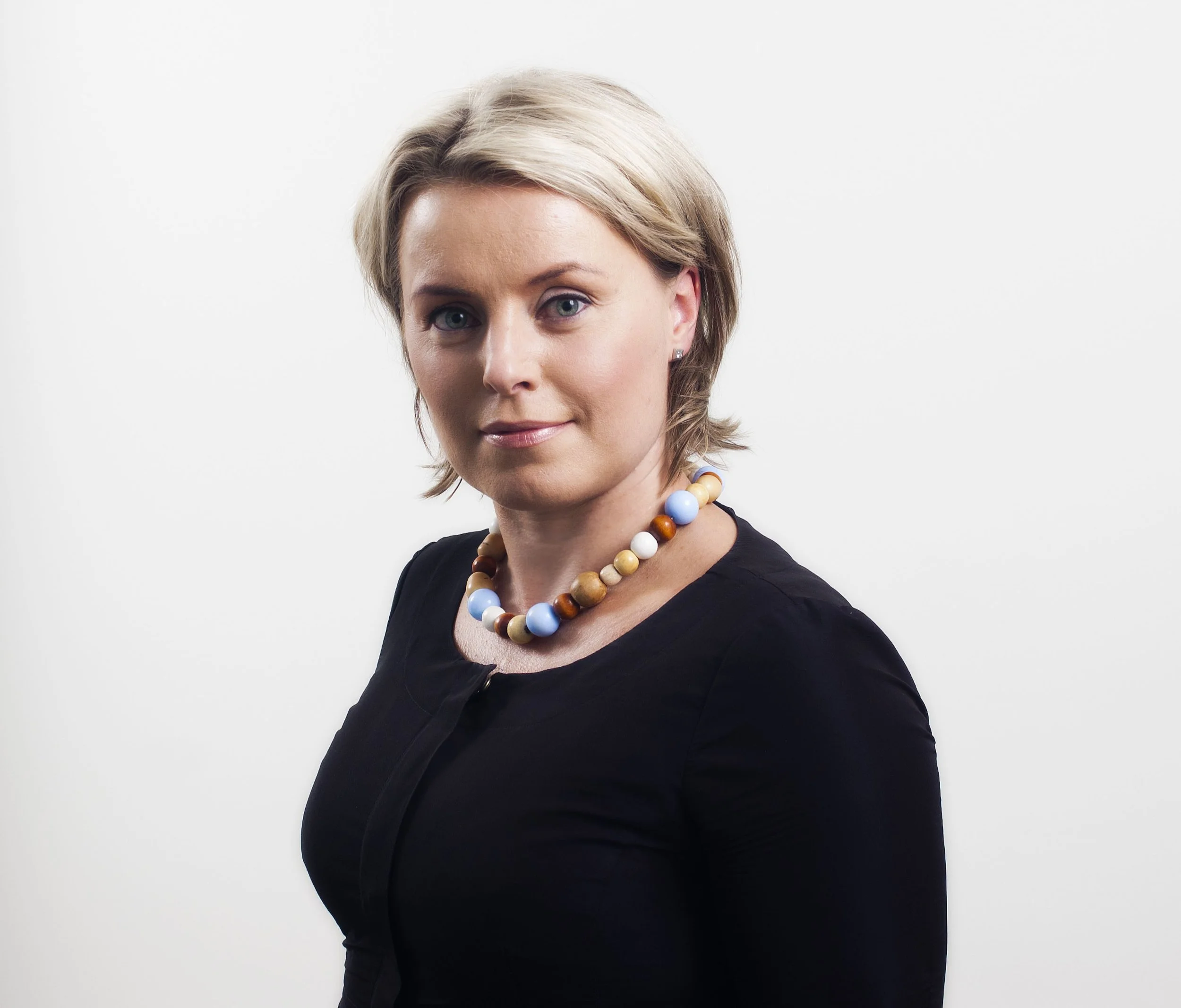 Portrait of a woman with short blond hair wearing a black top and a colorful bead necklace, standing against a plain white background.
