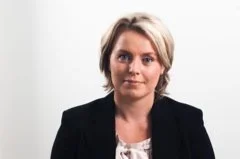 A woman with short blonde hair wearing a black blazer and white blouse, standing against a plain light background.