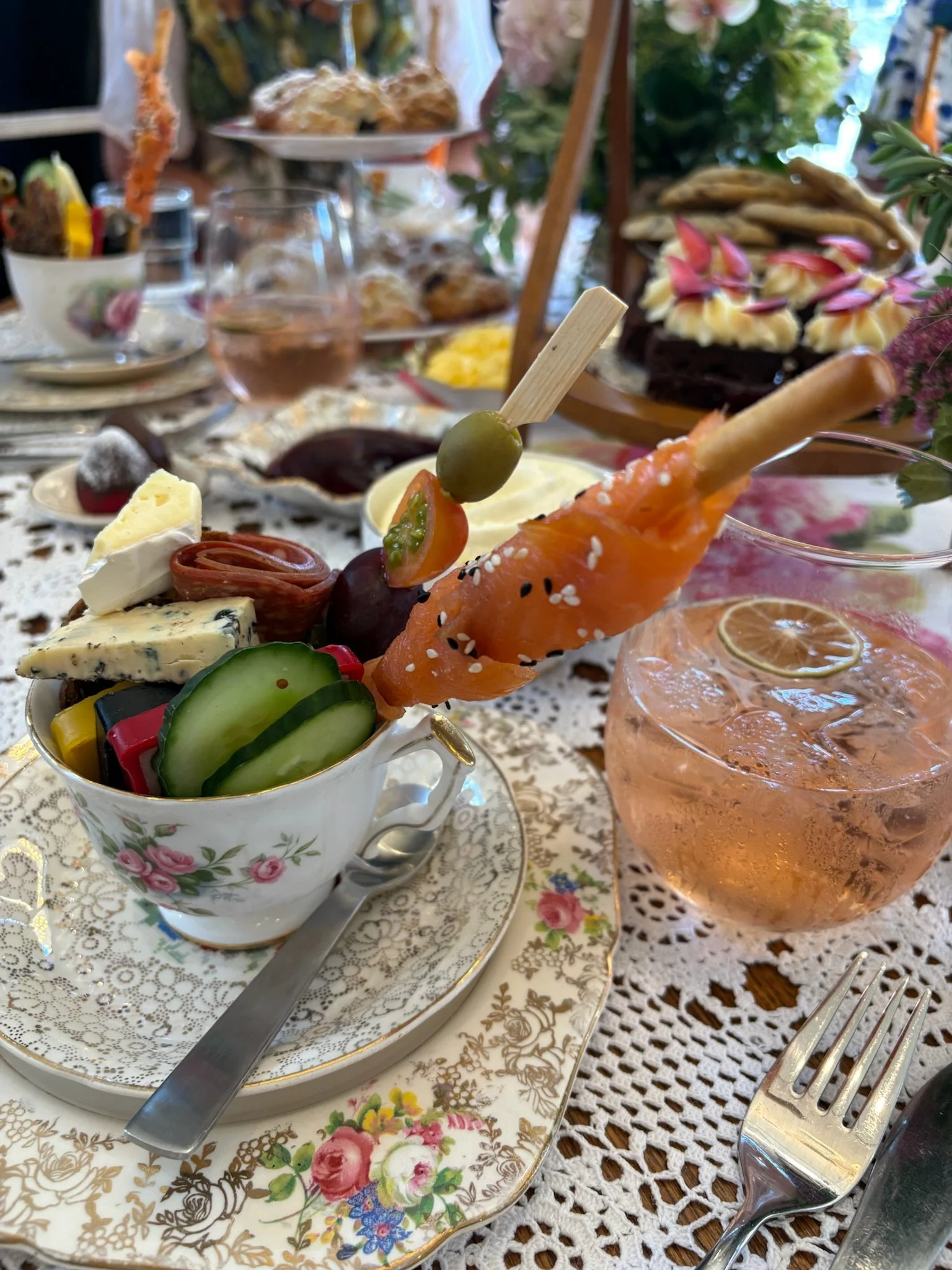 A festive table setting with a floral tea cup filled with sliced vegetables, cheeses, and meats, accompanied by a spoon and a fork on a lace tablecloth, with a glass of pink drink and a multi-layered tray of baked goods and desserts in the background.
