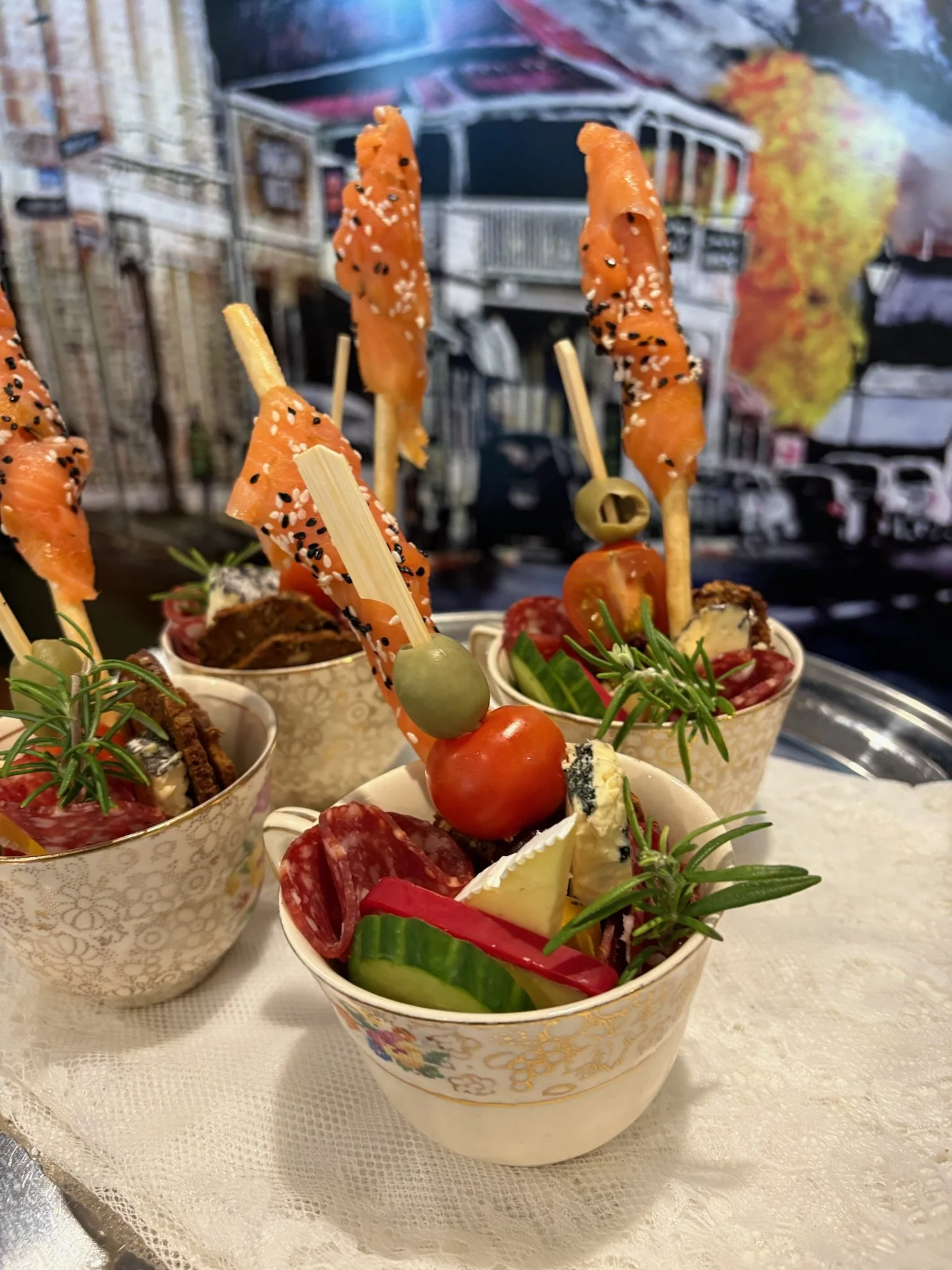 Assorted appetizers in decorative bowls, including skewered smoked salmon with sesame seeds, vegetable sticks, cheese, cherry tomatoes, olives, and cured meats, garnished with rosemary sprigs, on a lace tablecloth.