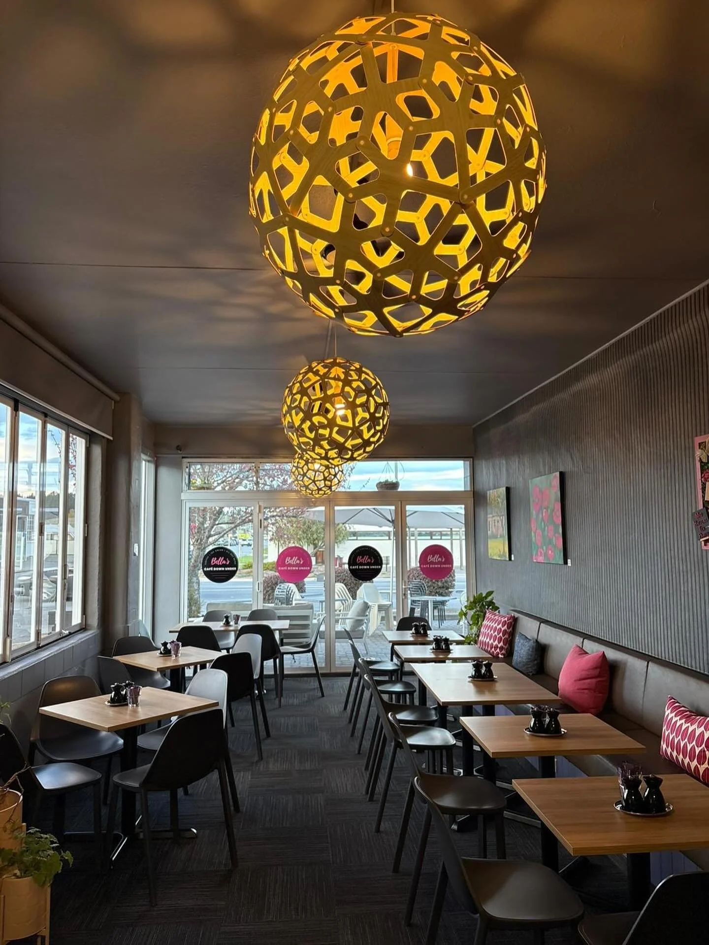 Interior of a cafe with wooden tables, dark grey chairs, pink and patterned cushions, large hanging pendant lights by a NZ designer, and a large window and glass door leading outside.
