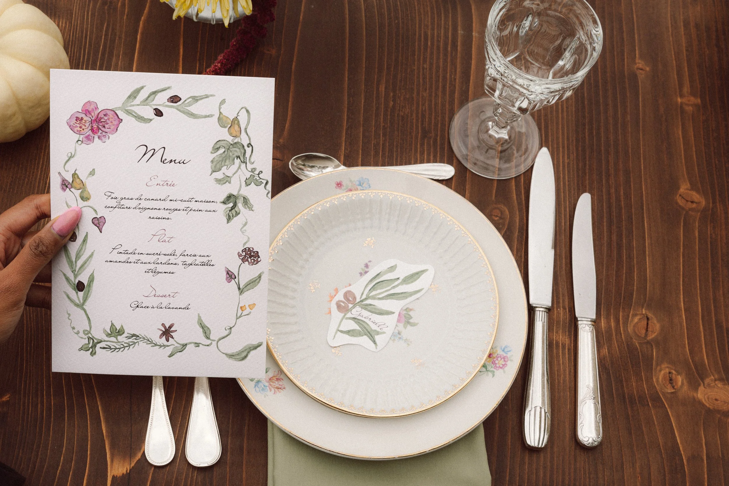 Une main tenant un menu de mariage au dessus d'une assiete. Illustration fleurie, feuilles, oliviers, couleurs autonmales