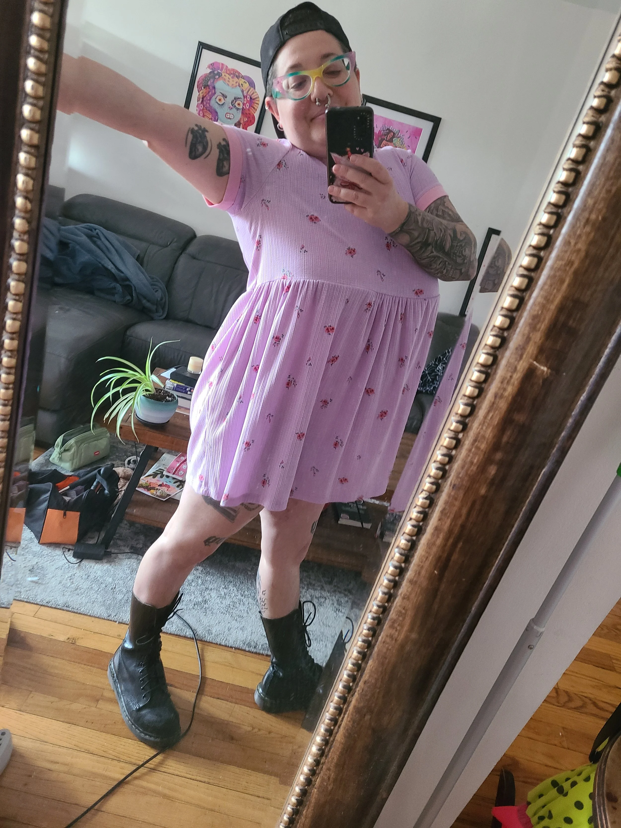 Founder of The Intuitive Assistant, Lina Akkerman, standing in front of a mirror, smiling with one arm outstretched. They are wearing a black baseball cap backwards, a light pink baby doll dress and black calf high doc martens
