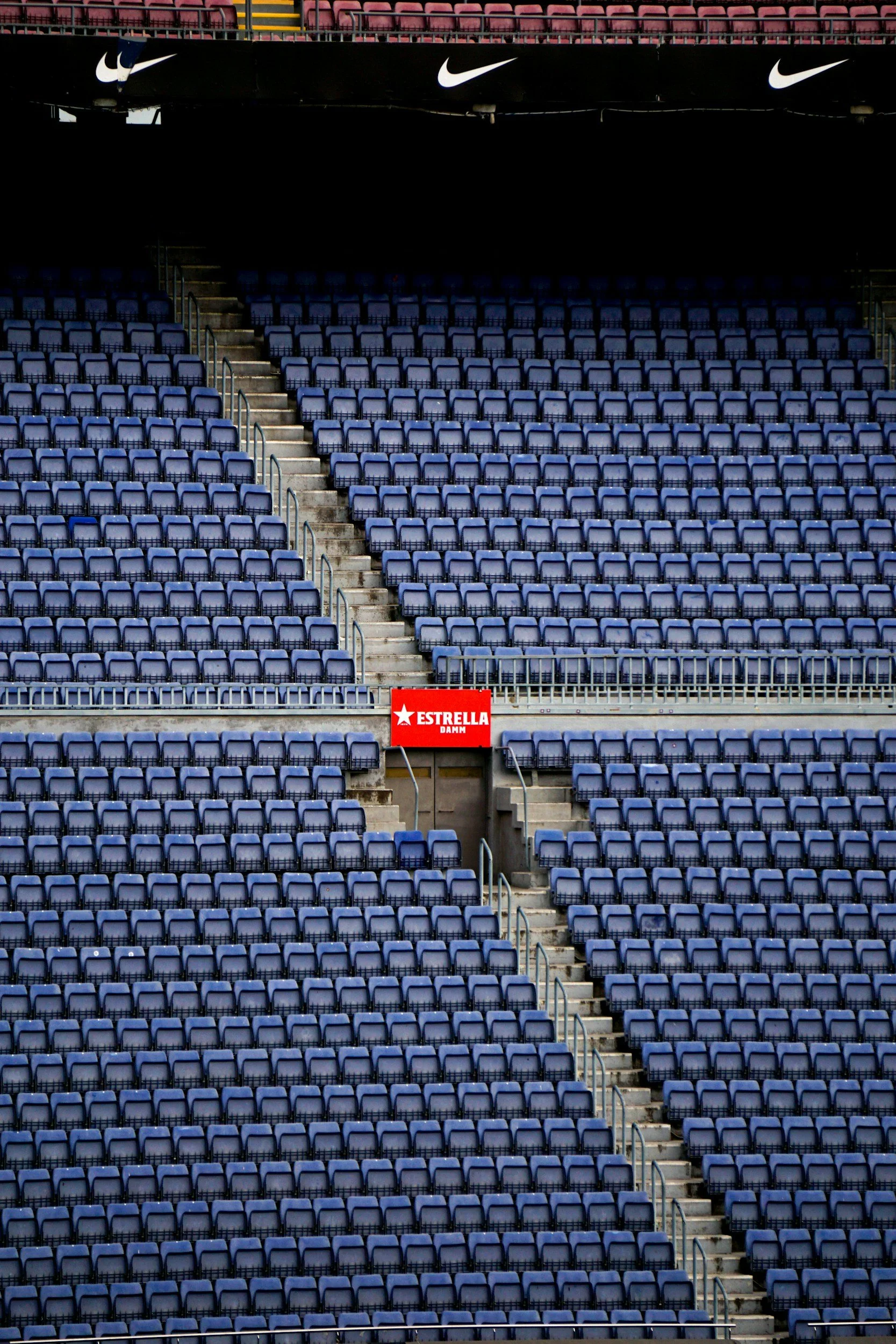 Empty stadium with blue seats, concrete stairs, and a red sign that reads "ESTRELLA DAMM" beneath Nike logos.