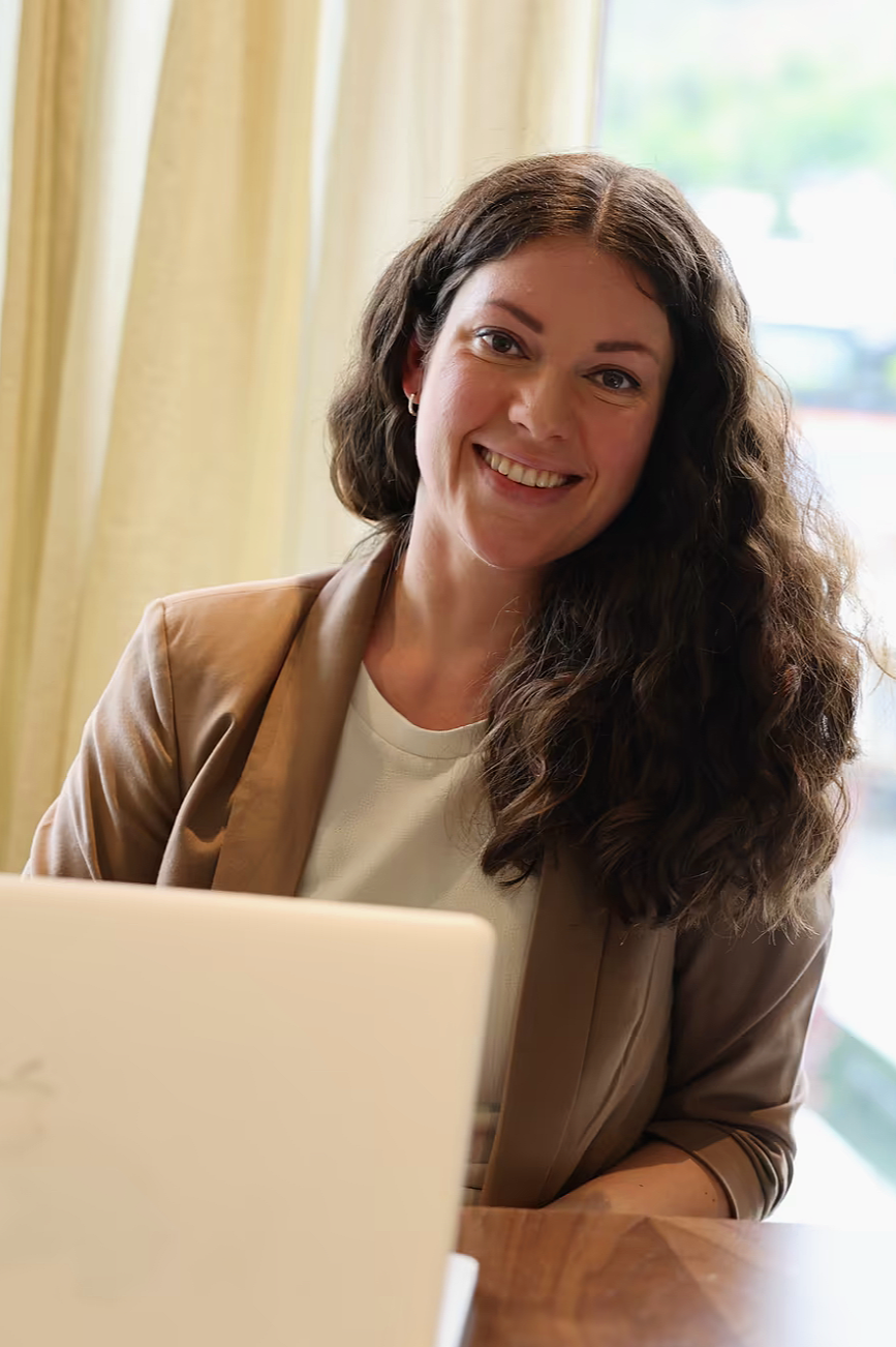 En kvinne med brunt, krøllete hår smiler og sitter foran en laptop i et rom med store vinduer og lyse gardiner.