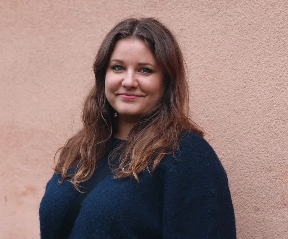 Et portrett av en smilende kvinne med langt brunt hår, iført en mørk blå genser, stående foran en lyserosa vegg.