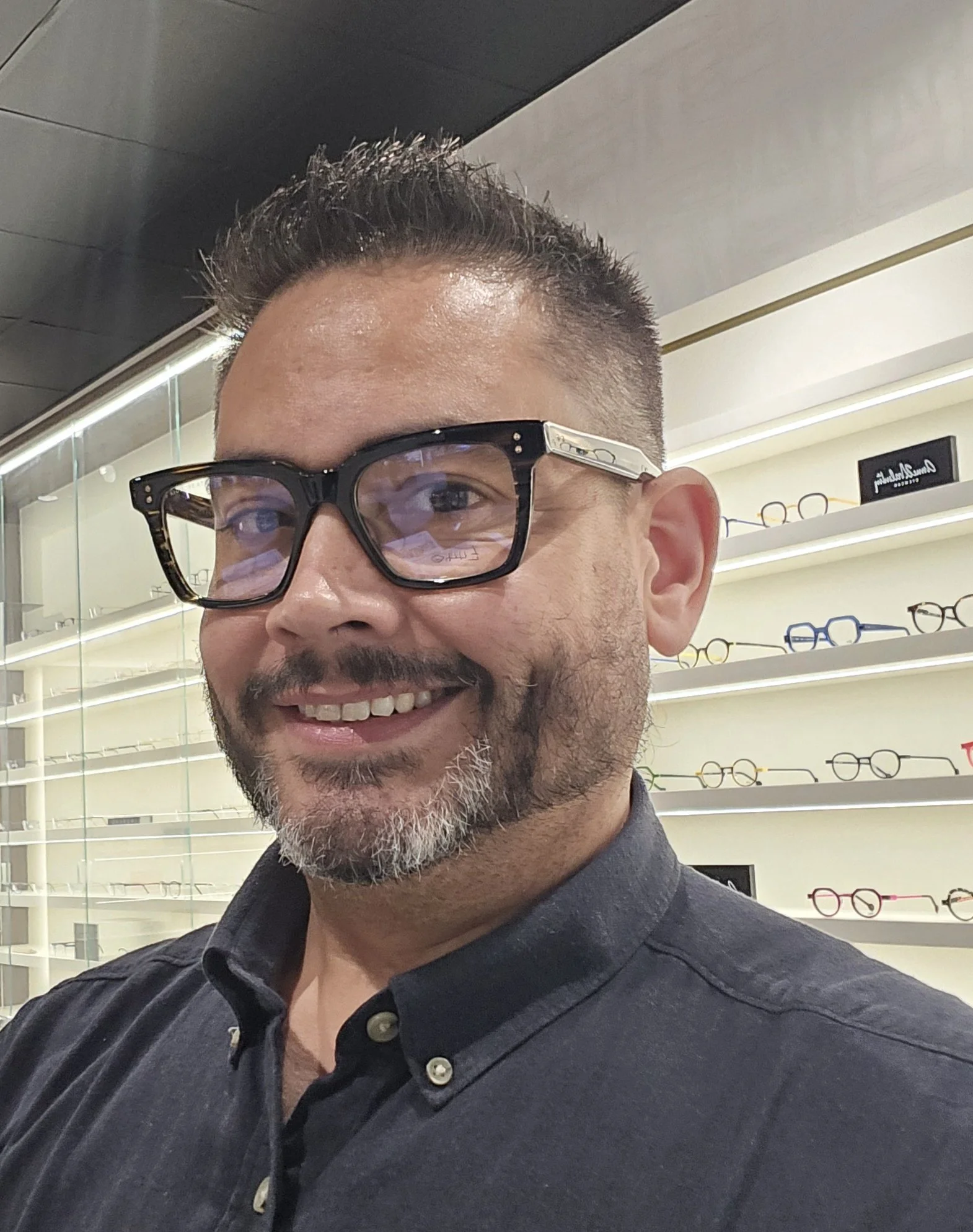 A man smiling in an eyewear store, wearing black glasses with decorative metal accents on the frame, in front of shelves displaying various eyeglasses.