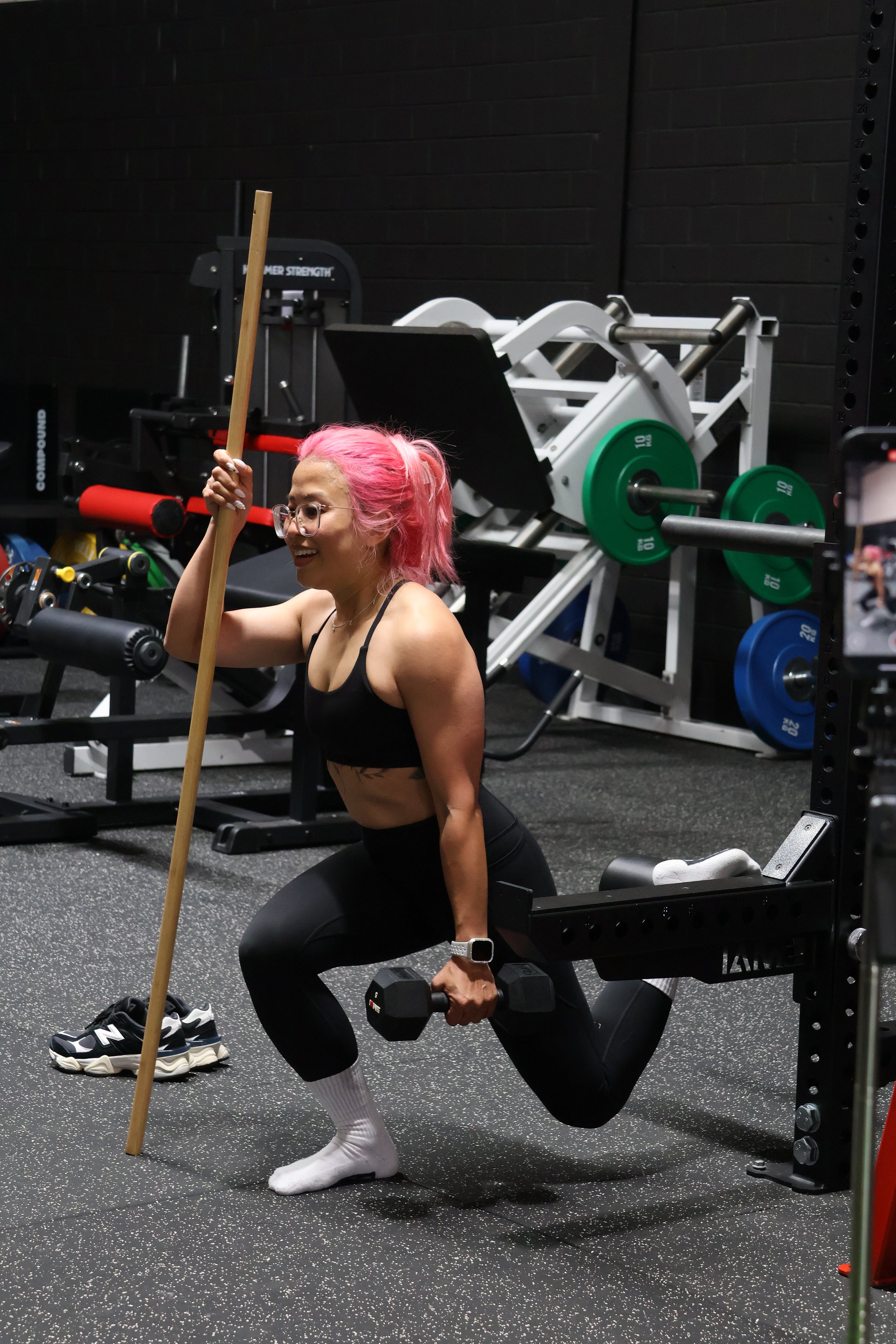 A woman with pink hair exercising in a gym, holding dumbbells and holding a broomstick model.