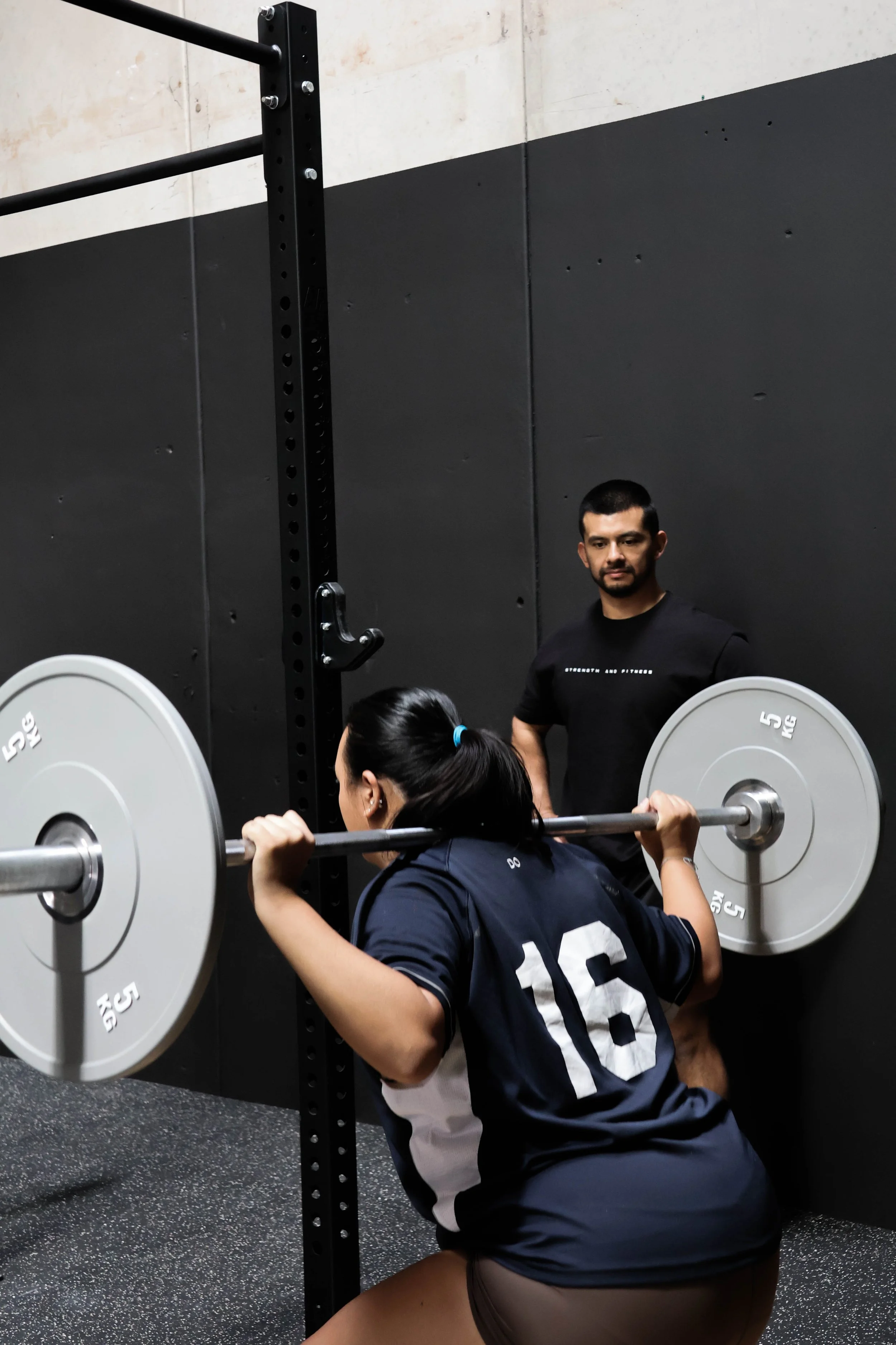 A woman performing a squat with a barbell on her shoulders, supervised by a man in a gym.