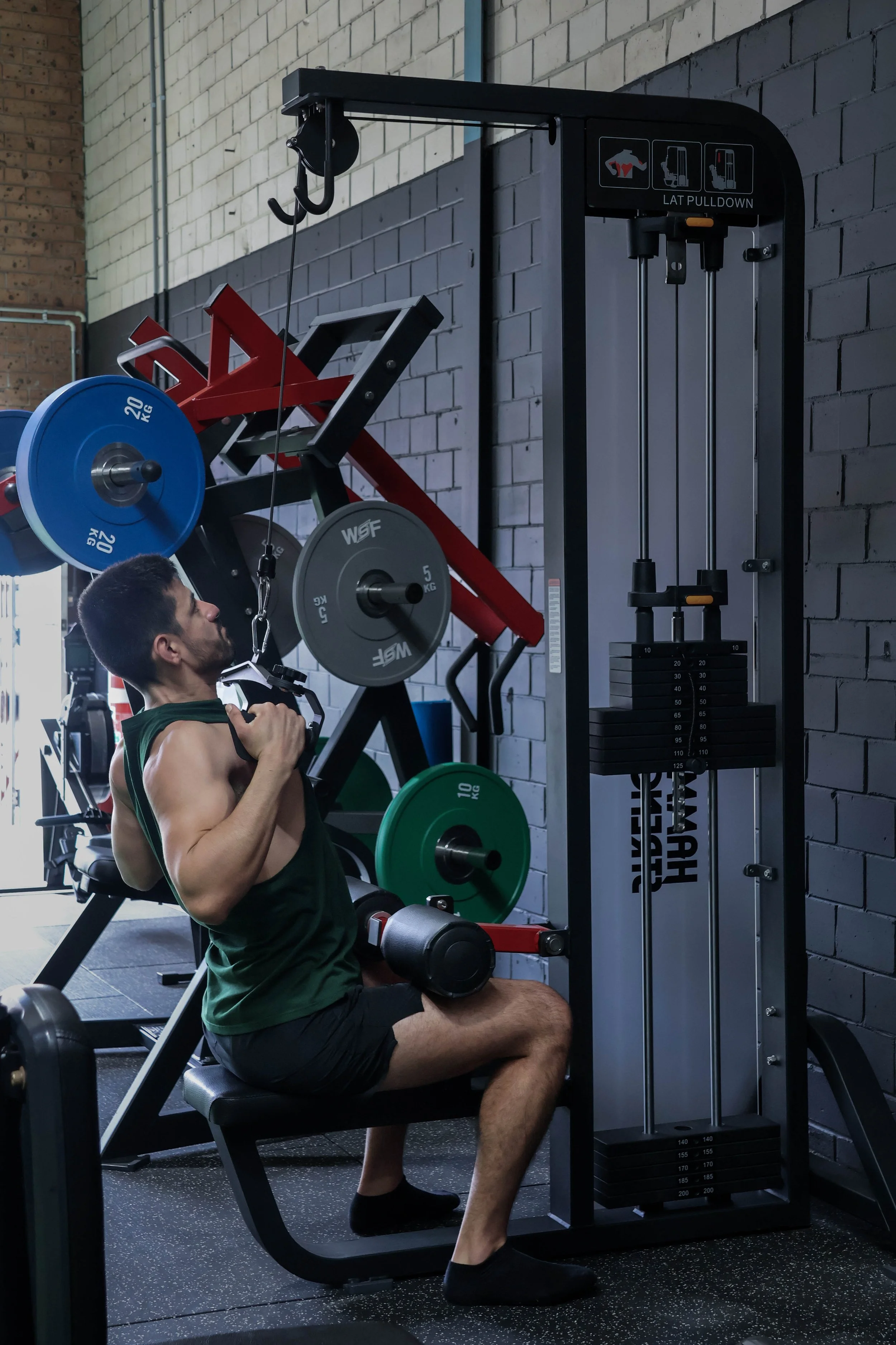 A man performs a shoulder press exercise on a seated machine at the gym, with weight plates and equipment in the background.