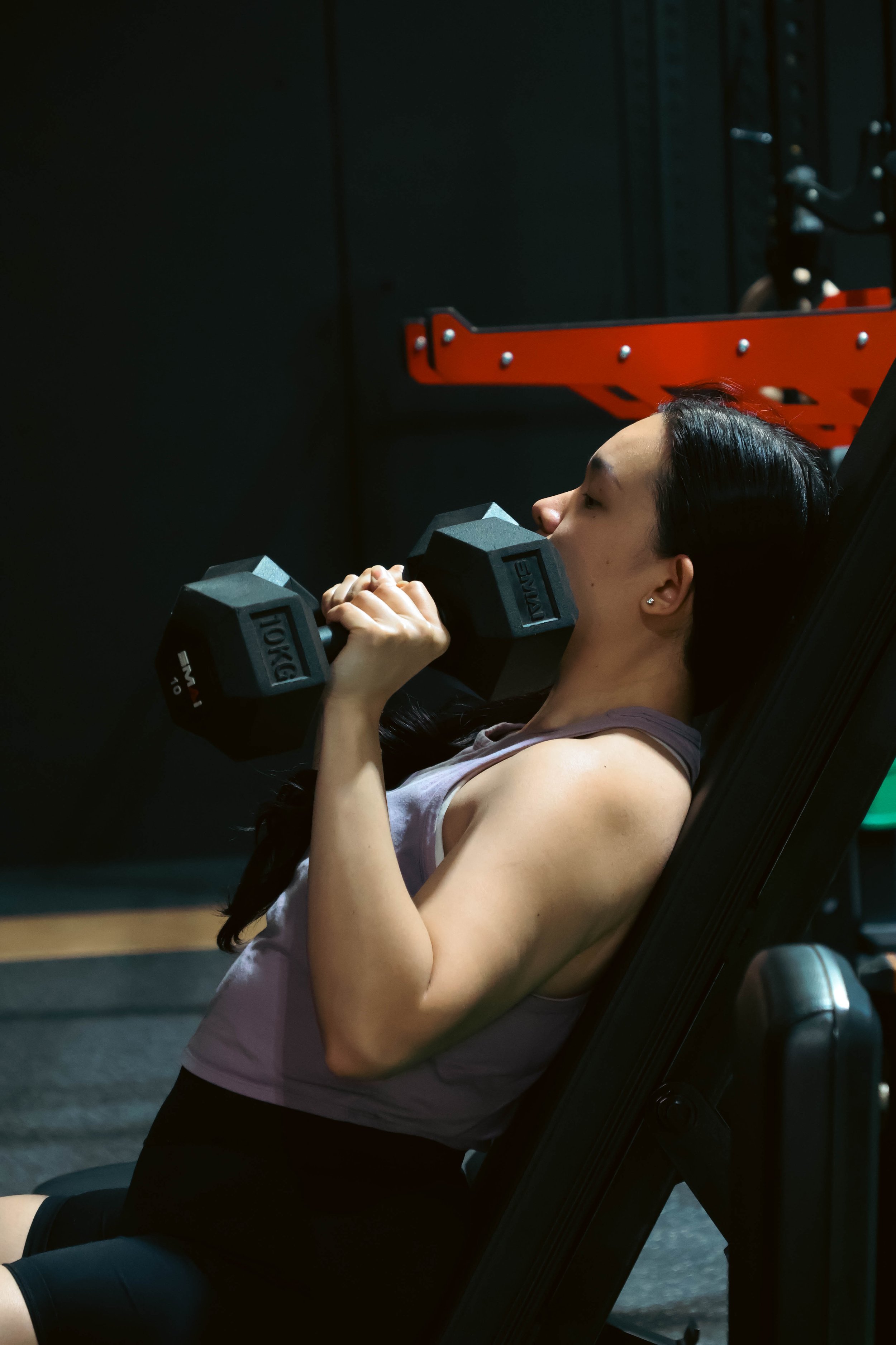 A woman lying on a workout bench lifting dumbbells in a gym.