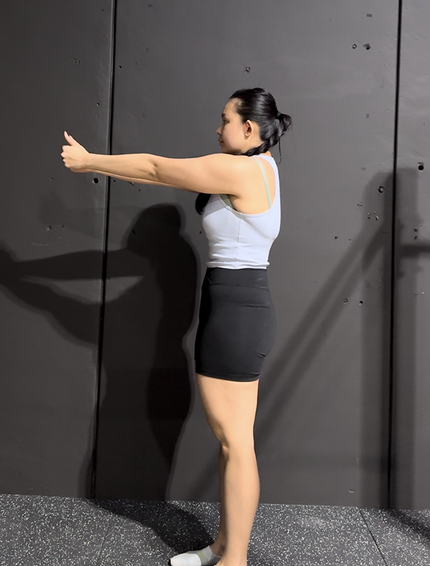 A woman performing a wall lean exercise against a black wall in a gym, wearing a white tank top and black shorts.