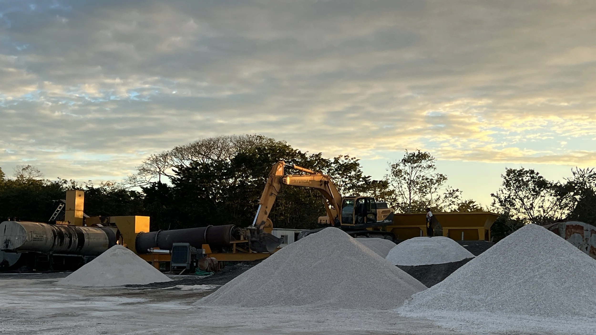 Construction site at sunset with piles of white gravel, a yellow excavator, and workers; trees and a cloudy sky in the background.