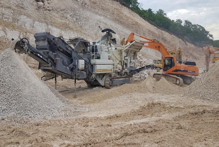 Construction site with a large excavator and a crushing machine on a dirt mound.
