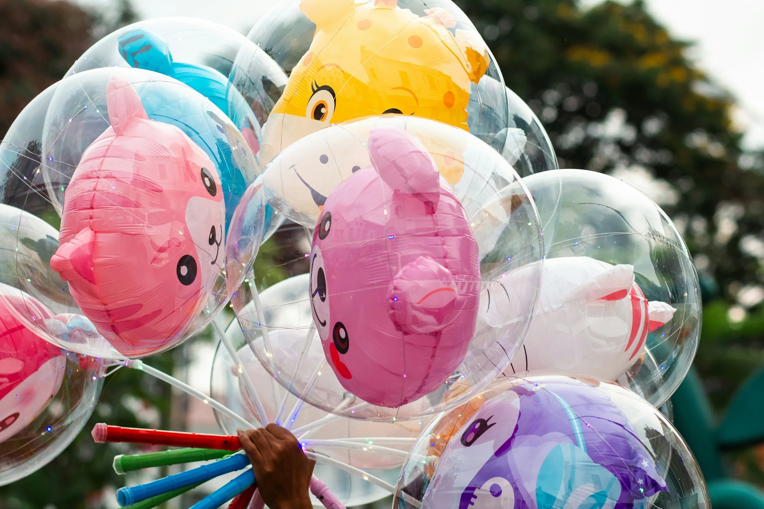 Colorful balloons with cute animal faces, including pigs, a bear, and a giraffe, held by multiple hands with rainbow-colored sticks, outdoors with trees in the background.