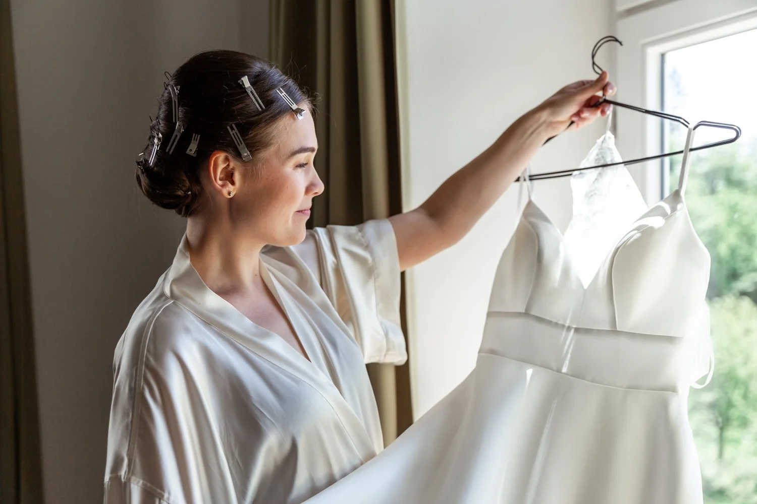 Eine lachende Frau mit Haarclips hält ein weißes Hochzeitskleid vor einem Fenster. Sie trägt ein cremefarbenes Satin-Shirt.