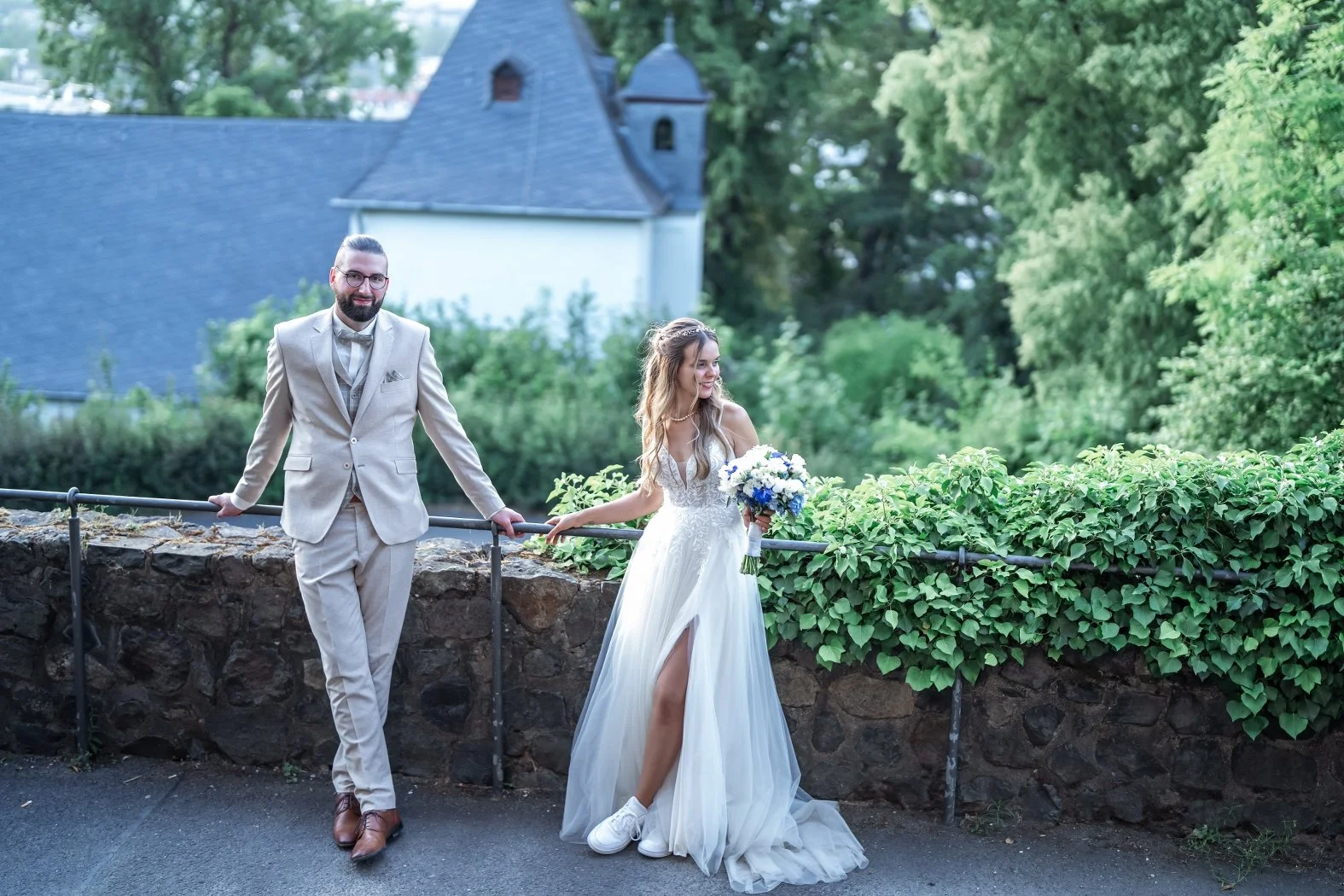 Ein Brautpaar steht neben einer Steinmauer im Freien, umgeben von grünen Bäumen. Die Frau trägt ein weißes Hochzeitskleid mit hoher Beinschlitze und hält einen Blumenstrauß, während der Mann einen cremefarbenen Anzug trägt.