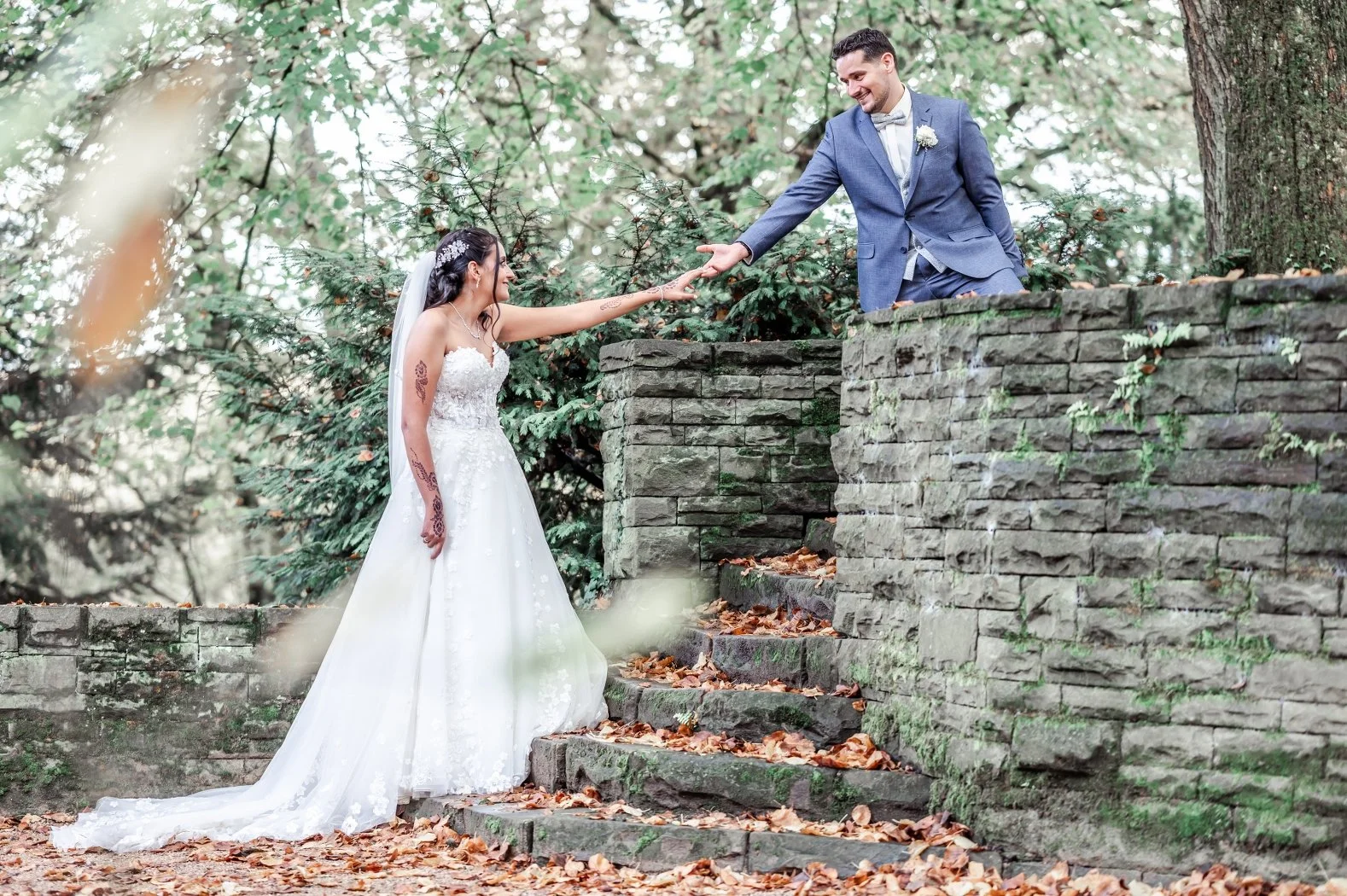 Ein Brautpaar bei einer Hochzeit im Freien, die Braut trägt ein weißes Hochzeitskleid mit Schleier, der Bräutigam trägt einen blauen Anzug mit Fliege, sie halten sich an den Händen und lächeln sich an, umgeben von Bäumen und Herbstlaub.