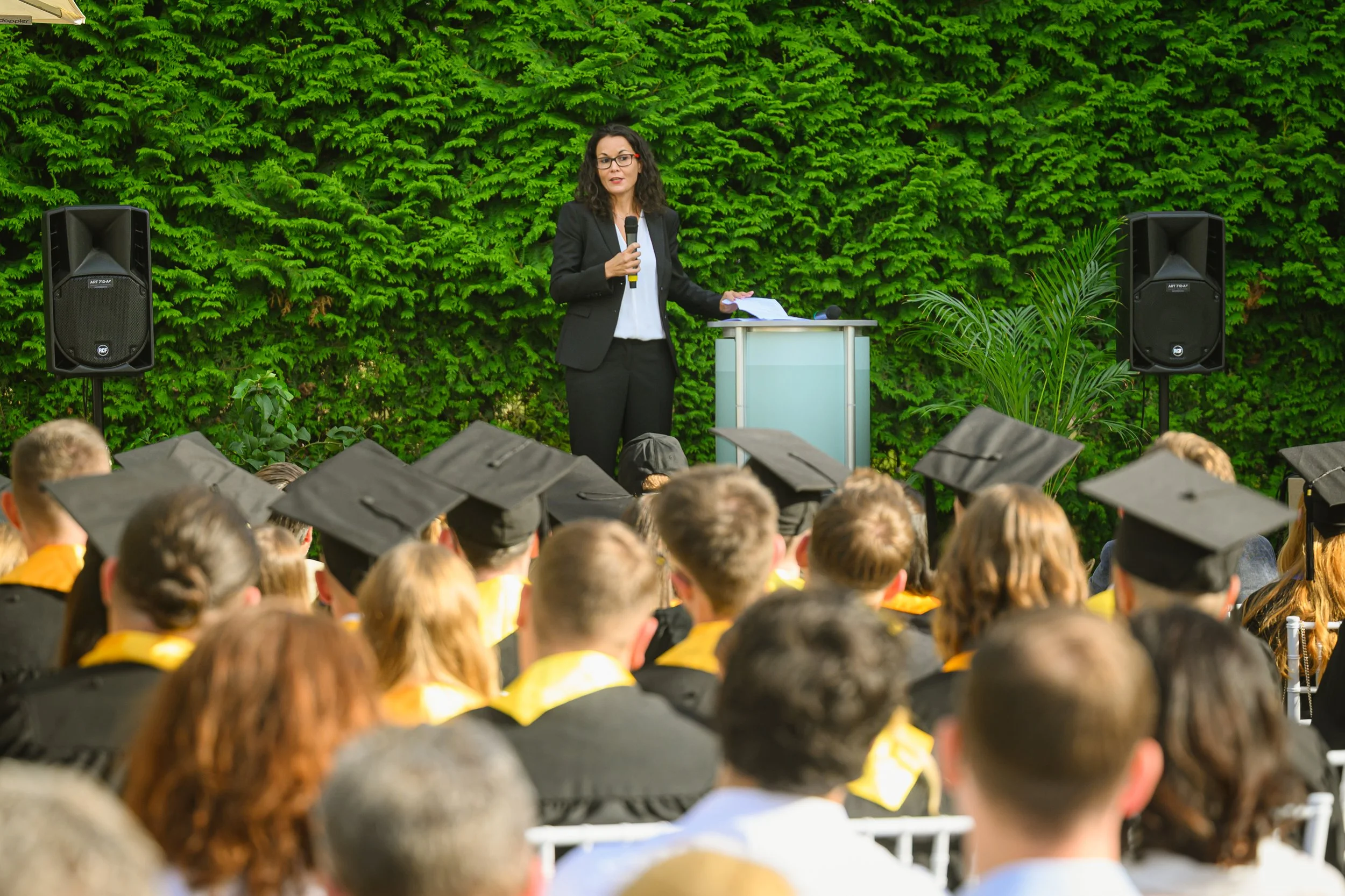 A woman in a black suit giving a speech at an outdoor graduation ceremony with graduates wearing caps and gowns seated in front of her, greenery in the background.