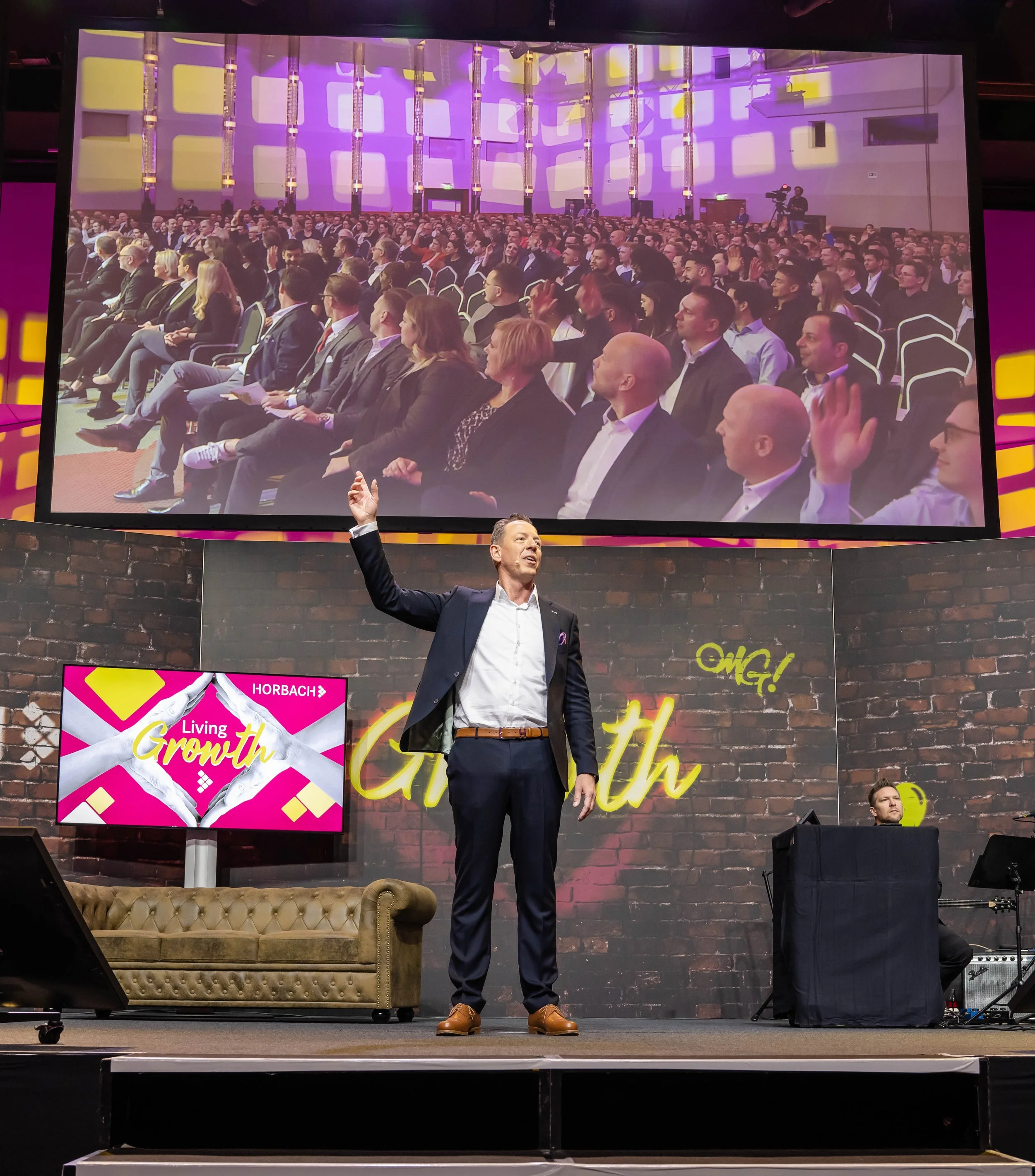 A man in a suit speaking on stage with a microphone, in front of a large screen displaying an audience of people seated at an event called Living Growth, with a brick wall backdrop and a small live band setup.