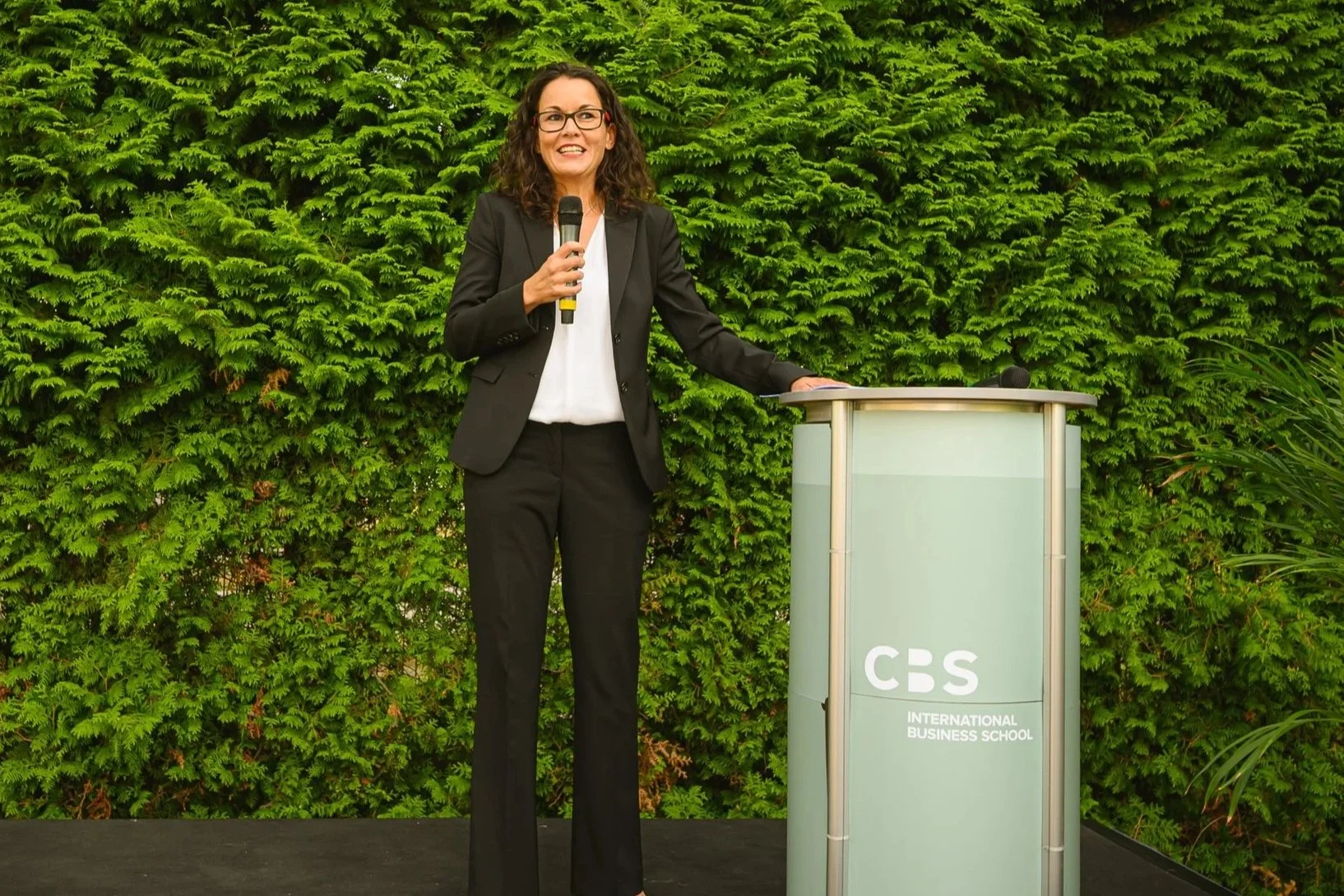 A woman in a black suit and glasses speaking at a podium with a CBS International Business School logo, outdoors with a lush green hedge background.