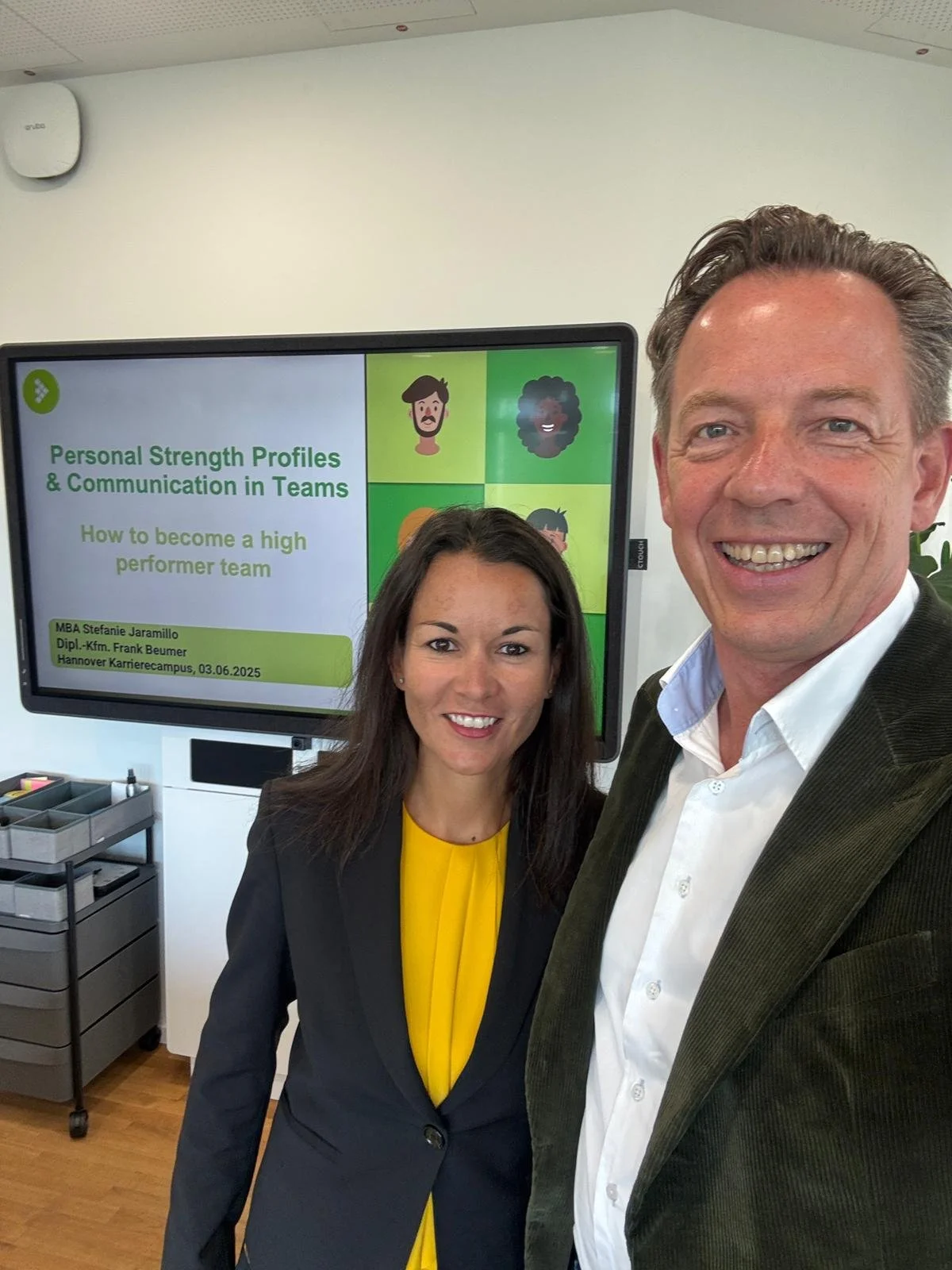 A man and woman taking a selfie in front of a presentation screen at a professional event. The screen displays a slide titled 'Personal Strength Profiles & Communication in Teams,' with the subtitle 'How to become a high performer team,' and details about a speaker, MBA Stefanie Jaramillo.