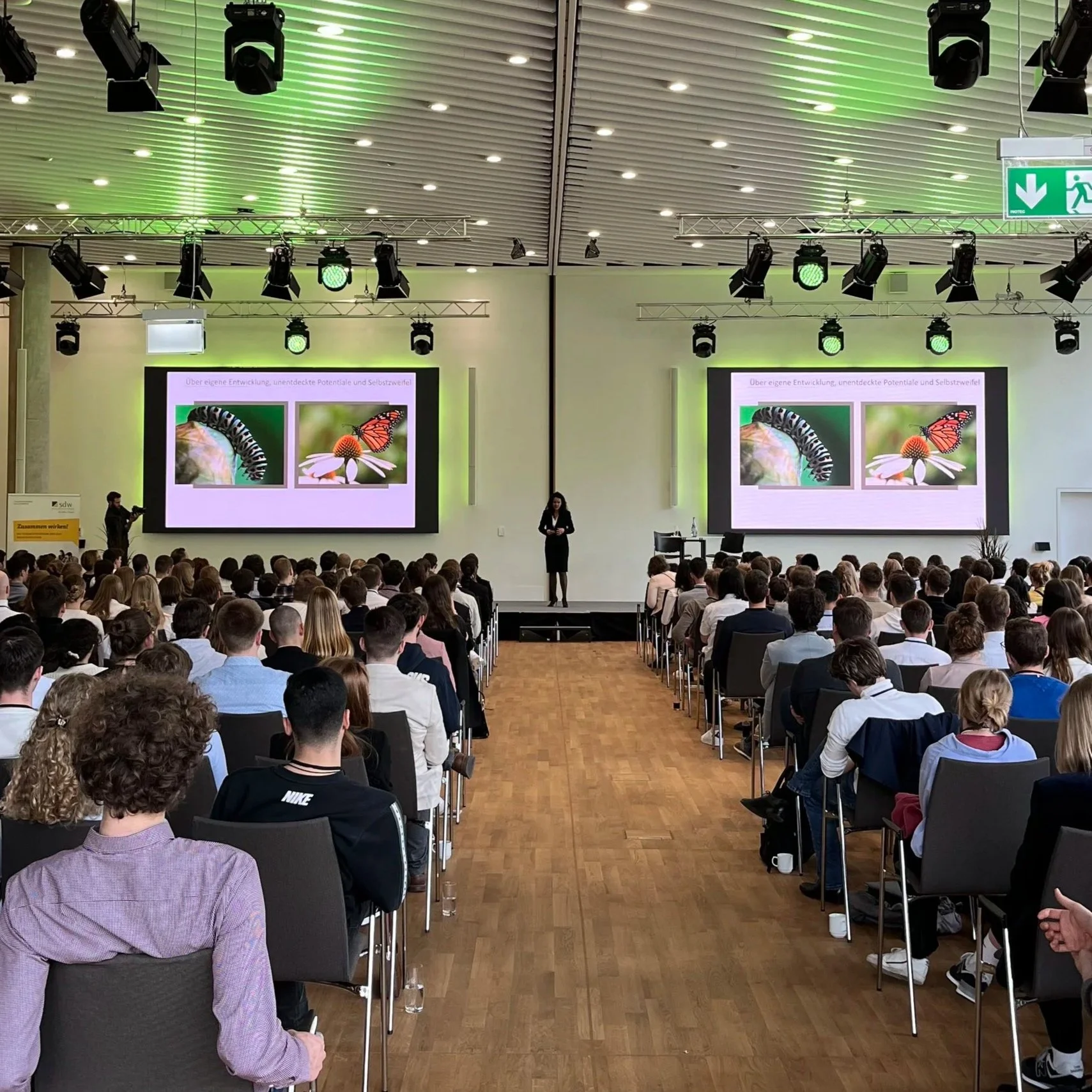 A large conference room filled with attendees watching a presentation. The presenter stands on stage with two screens behind showing images of a caterpillar and a butterfly. The room has a high ceiling with spotlighting and green lighting accents.