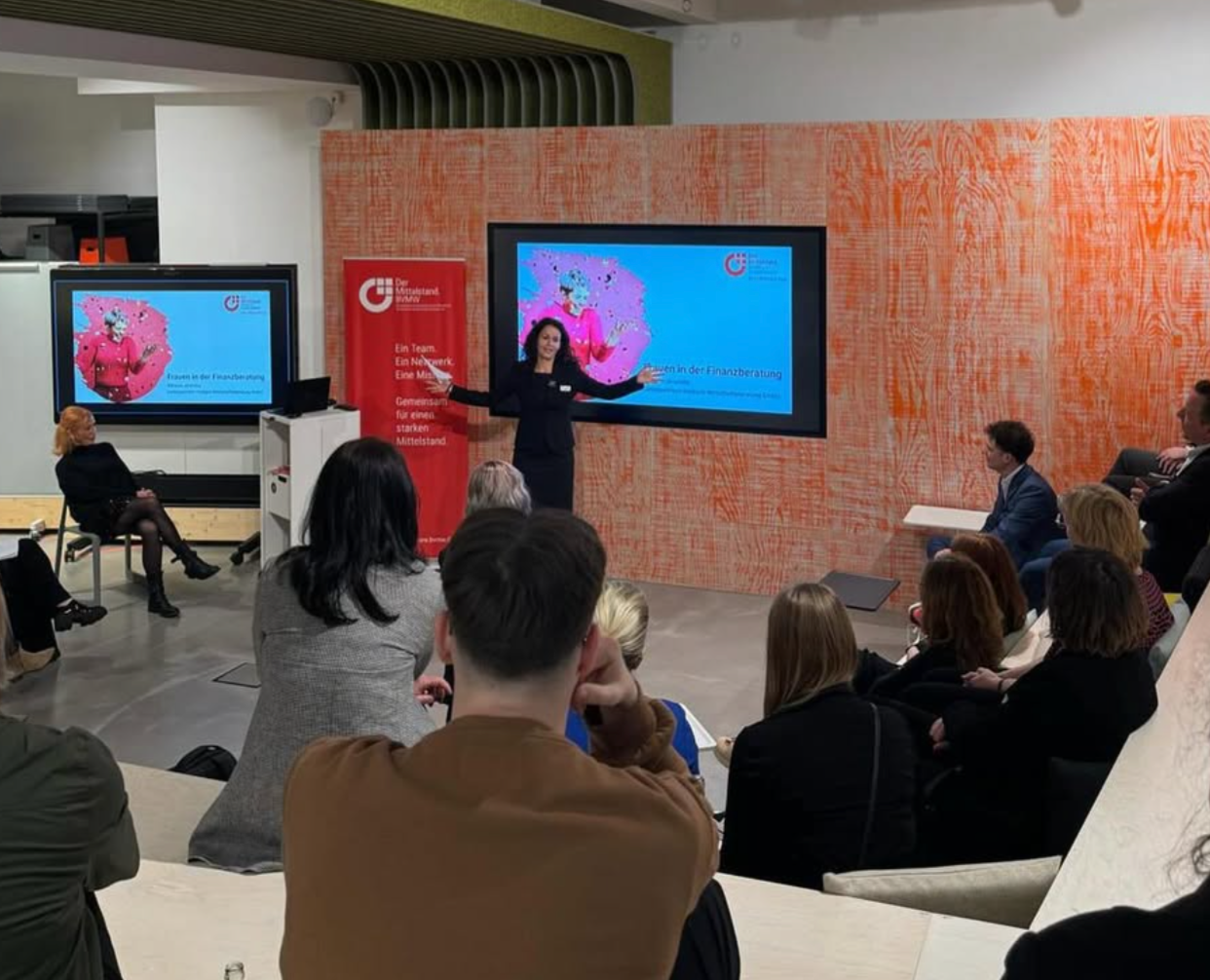 A woman giving a presentation in front of a large screen at a seminar or conference, with attendees seated and listening.