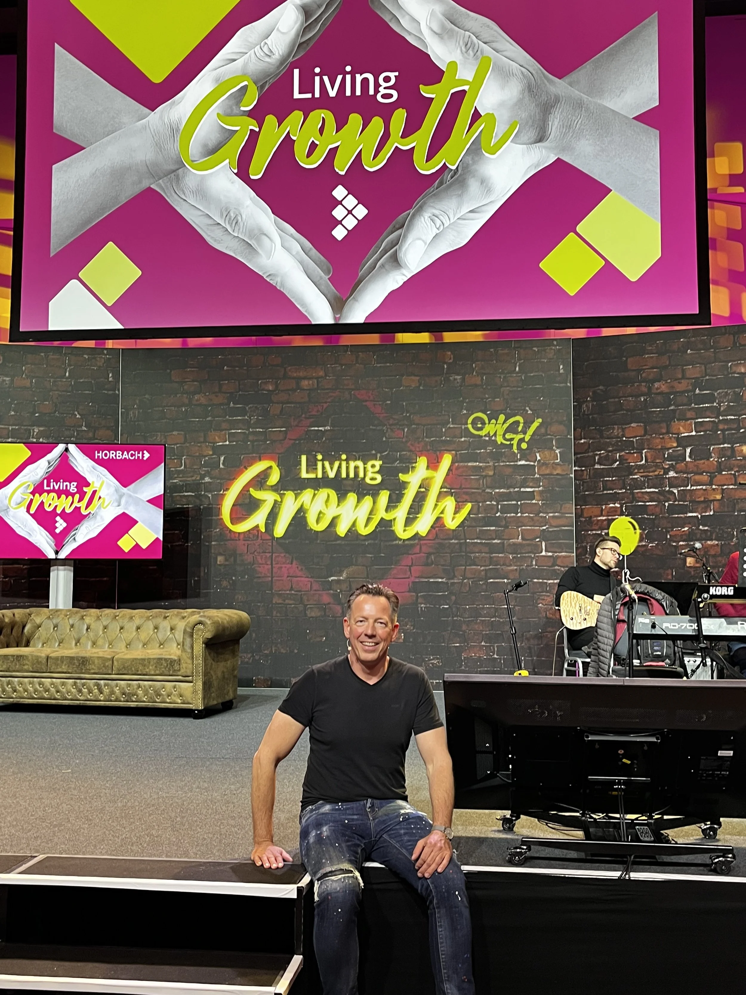 A man smiling and sitting on a stage at a 'Living Growth' event with a brick wall background. Two screens display the event name, and two people playing musical instruments are visible in the background.