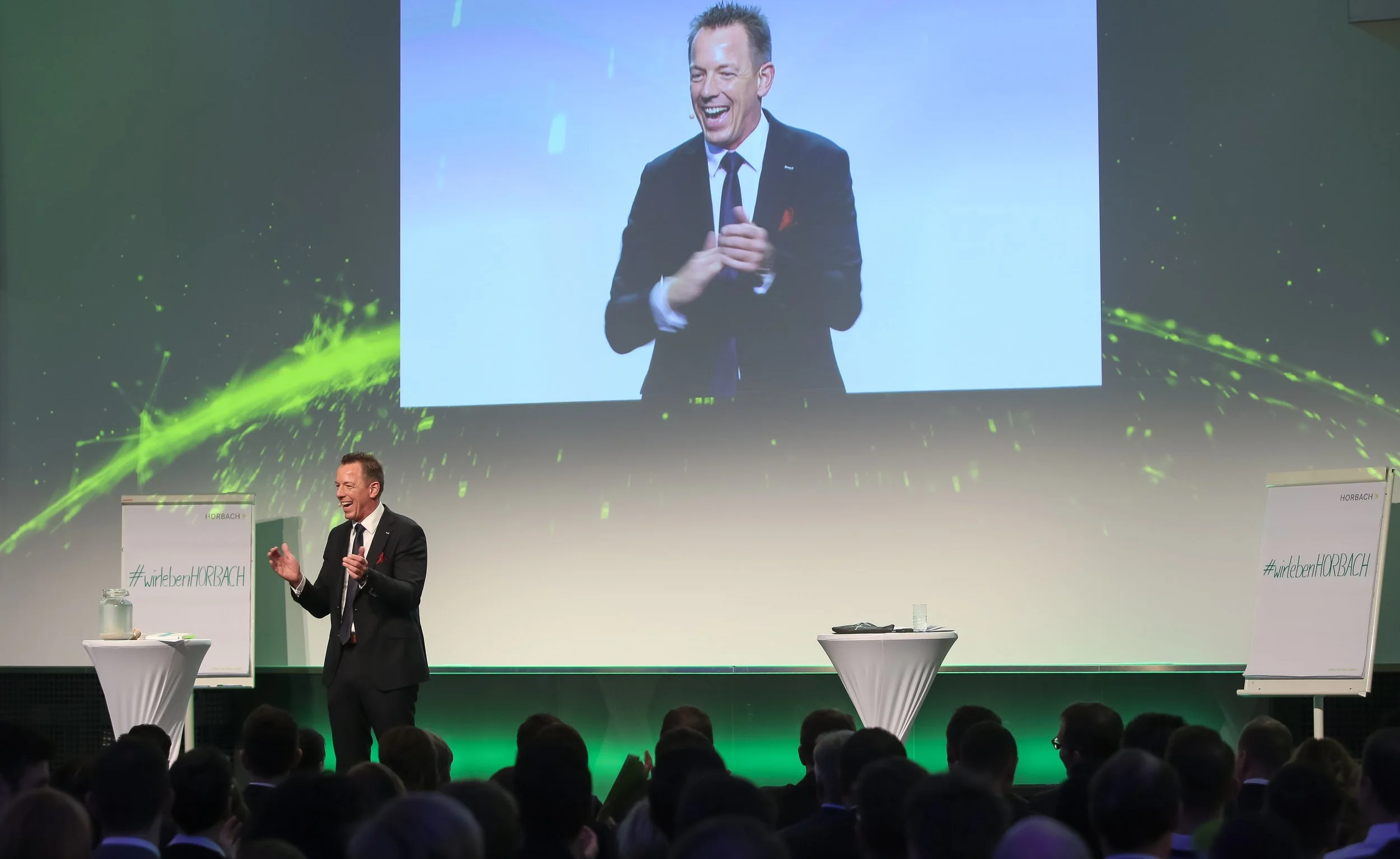 A man in a dark suit with a white shirt and a dark tie is standing on stage, smiling and clapping in front of an audience. A large screen behind him displays his image, and there are two whiteboards with text, one on each side of the stage.